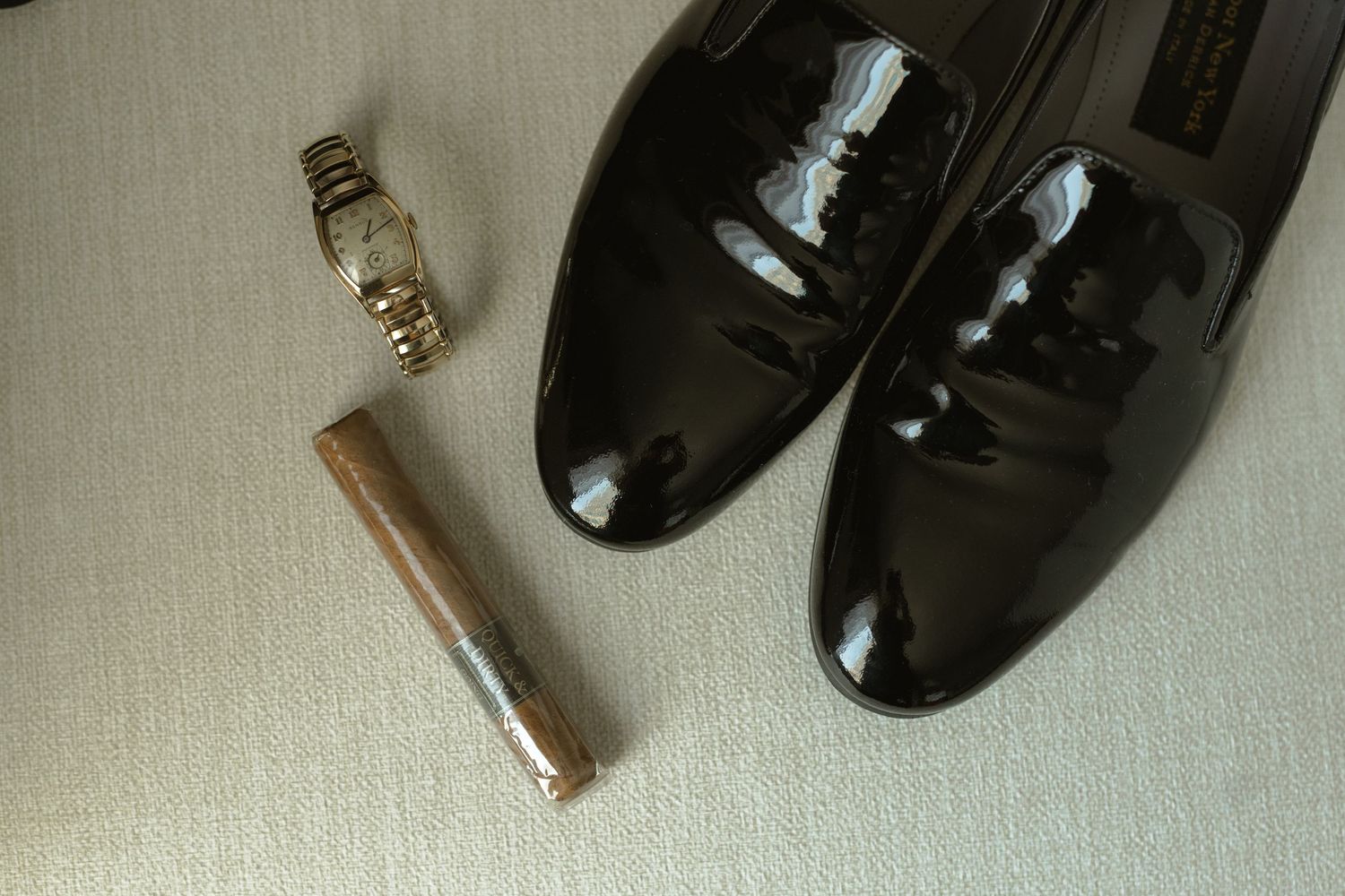 Wedding accessories including black patent leather shoes, rings and lipstick on light fabric.