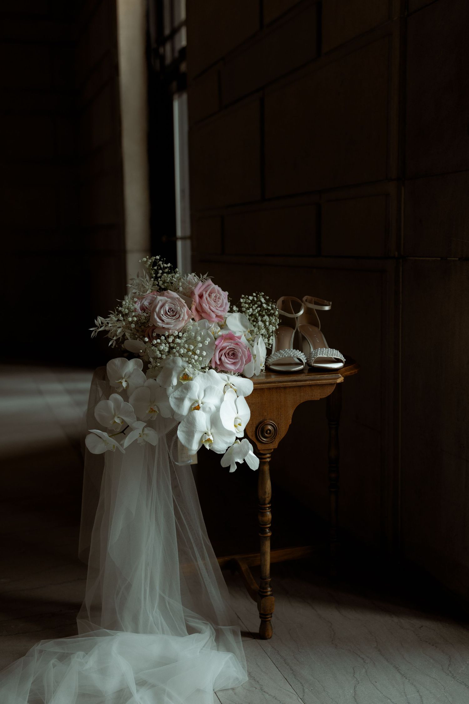 A delicate bridal veil and bouquet of pink roses and white orchids rest on an antique wooden table in moody lighting.
