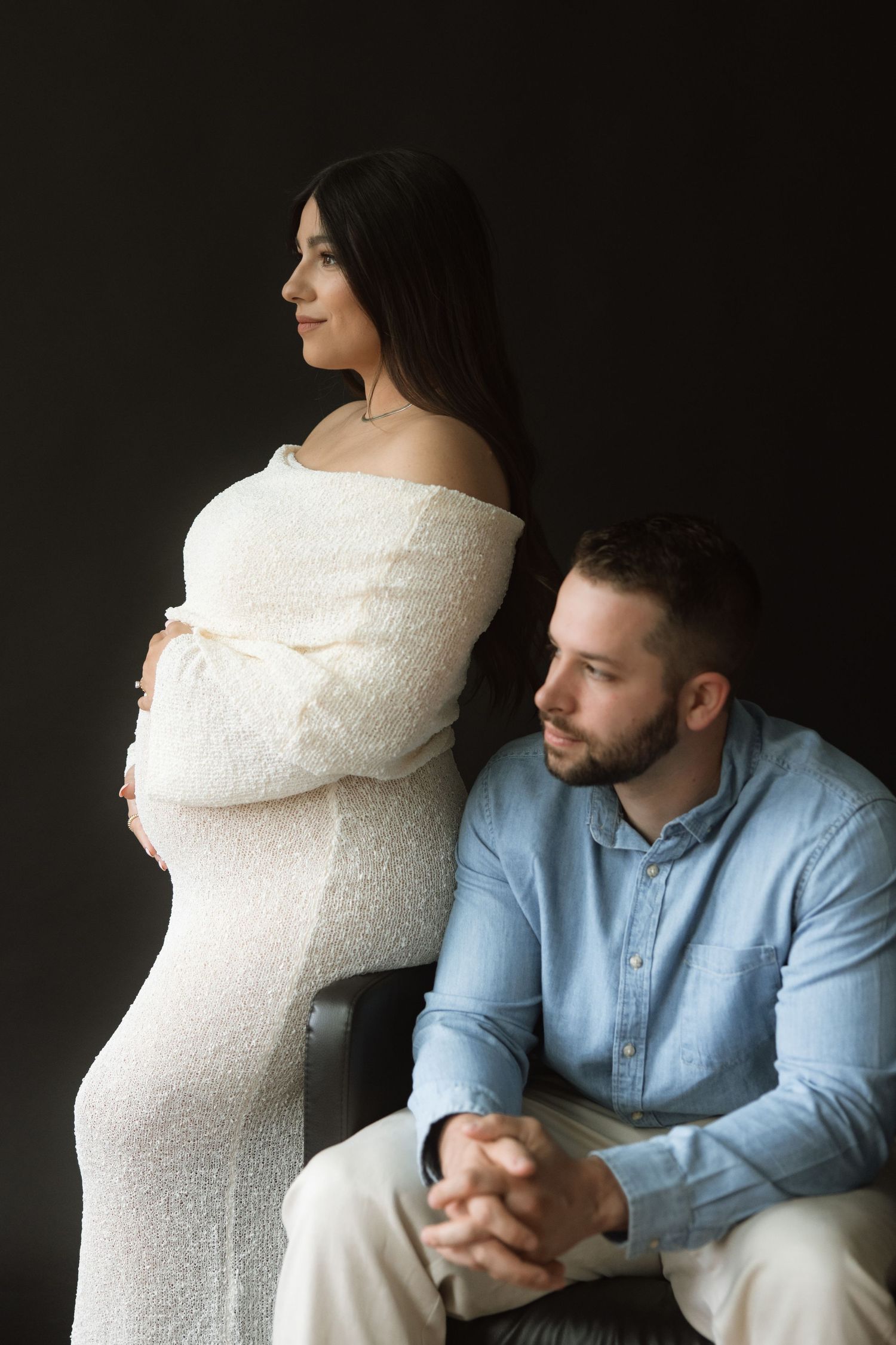 An intimate maternity portrait shows an expectant couple in formal attire against a dark backdrop.