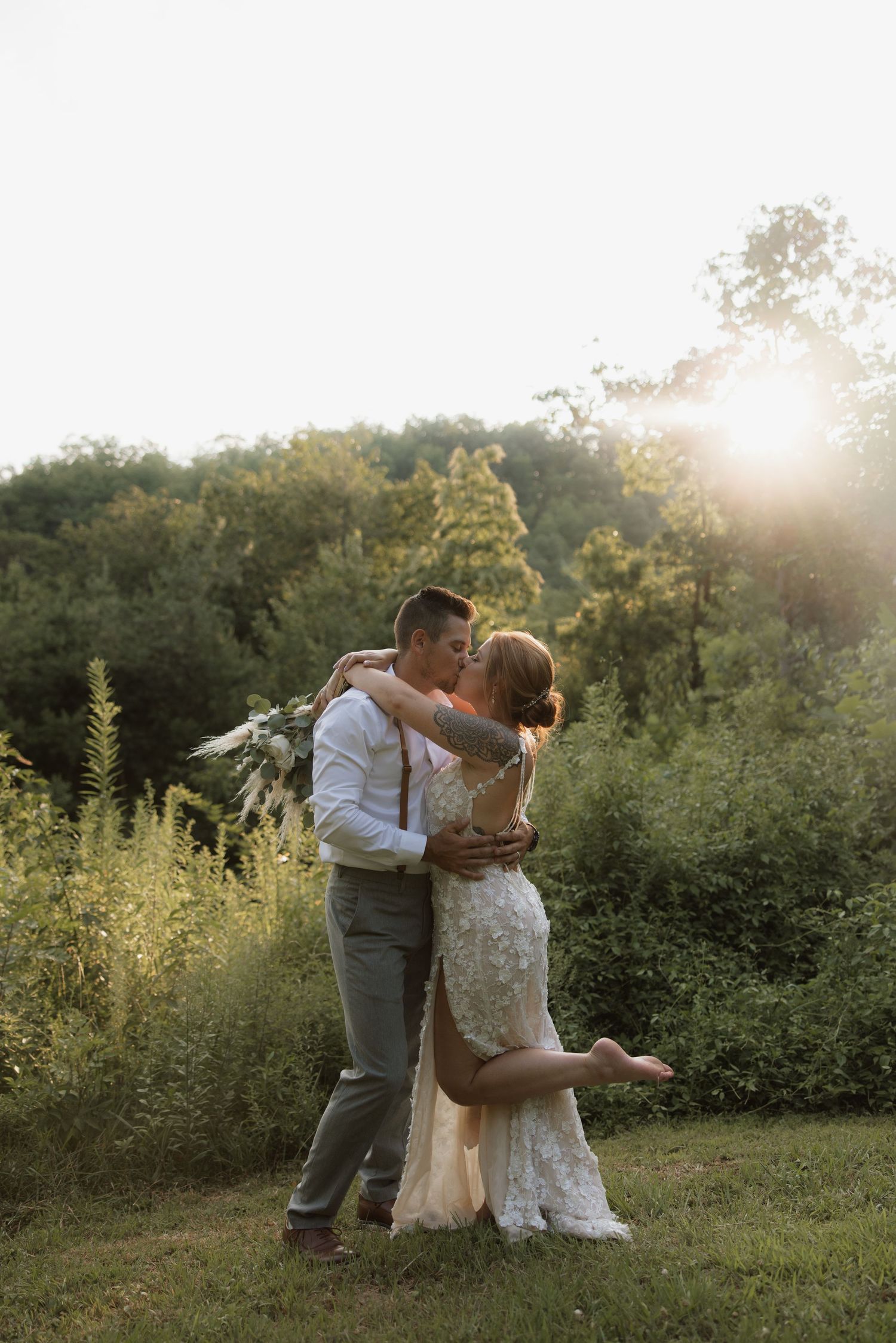 Couple embraces in a romantic kiss on a grassy hill during golden hour with sunlit trees in the background.