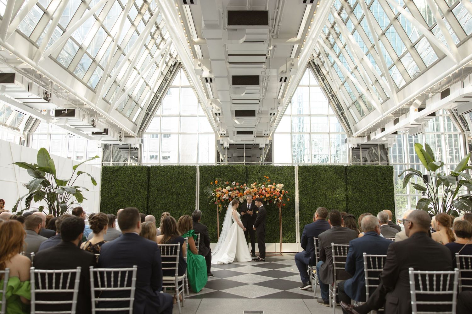Modern greenhouse wedding ceremony with white chairs and lush floral arrangements against hedge walls.