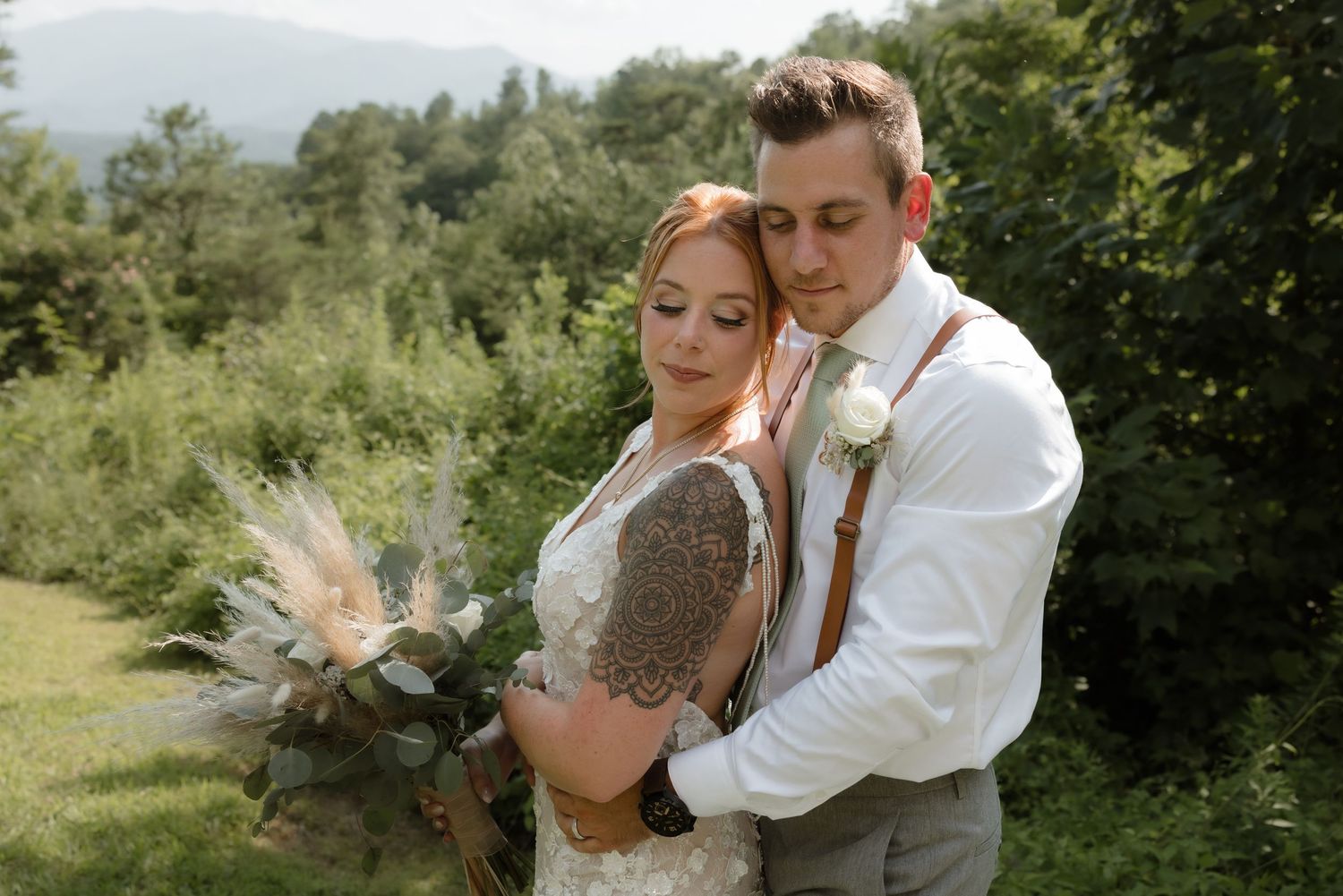 A romantic couple embraces outdoors on their wedding day surrounded by green mountain scenery.