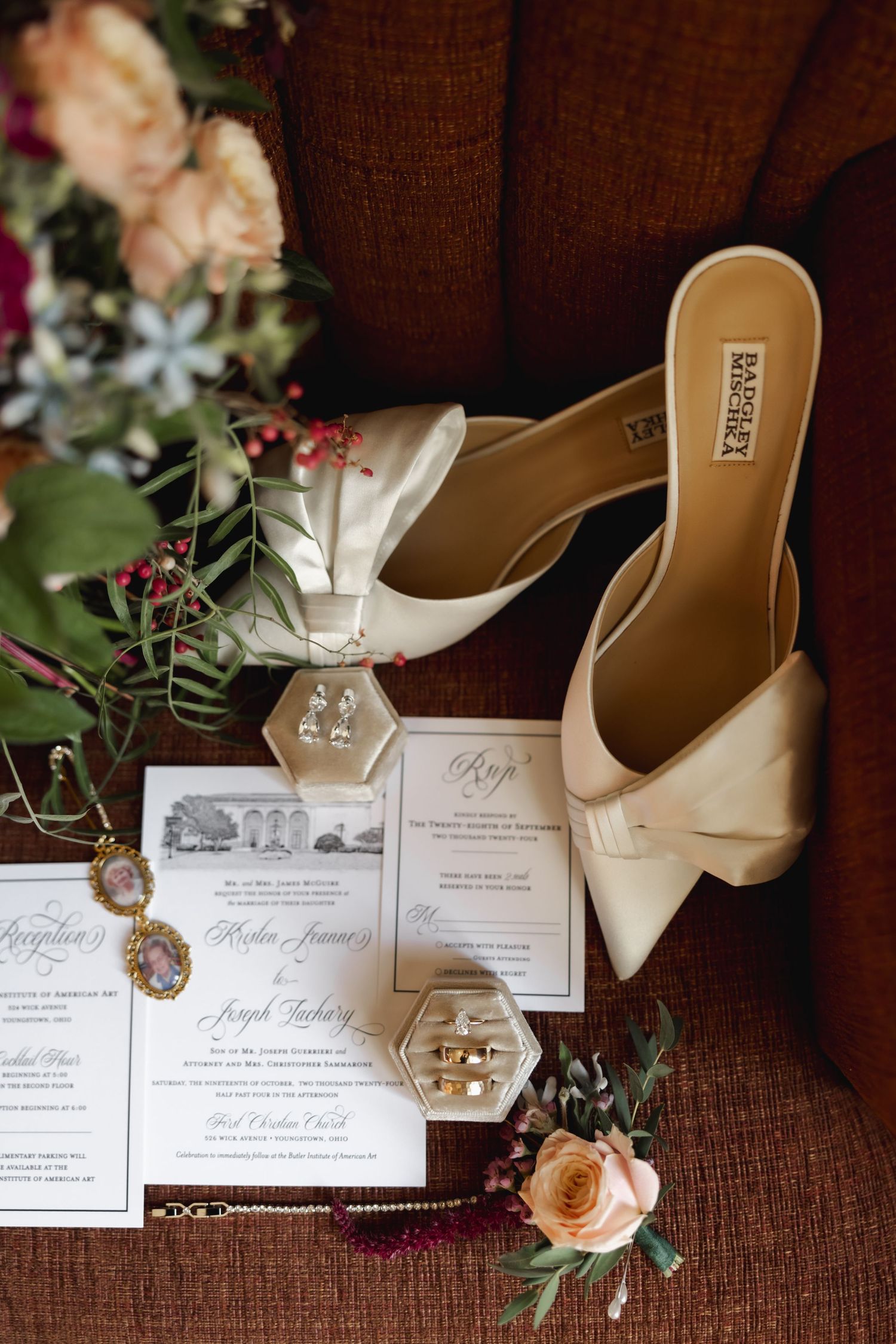 Wedding invitations and cream-colored heels arranged with delicate peach roses and greenery on brown textured fabric.