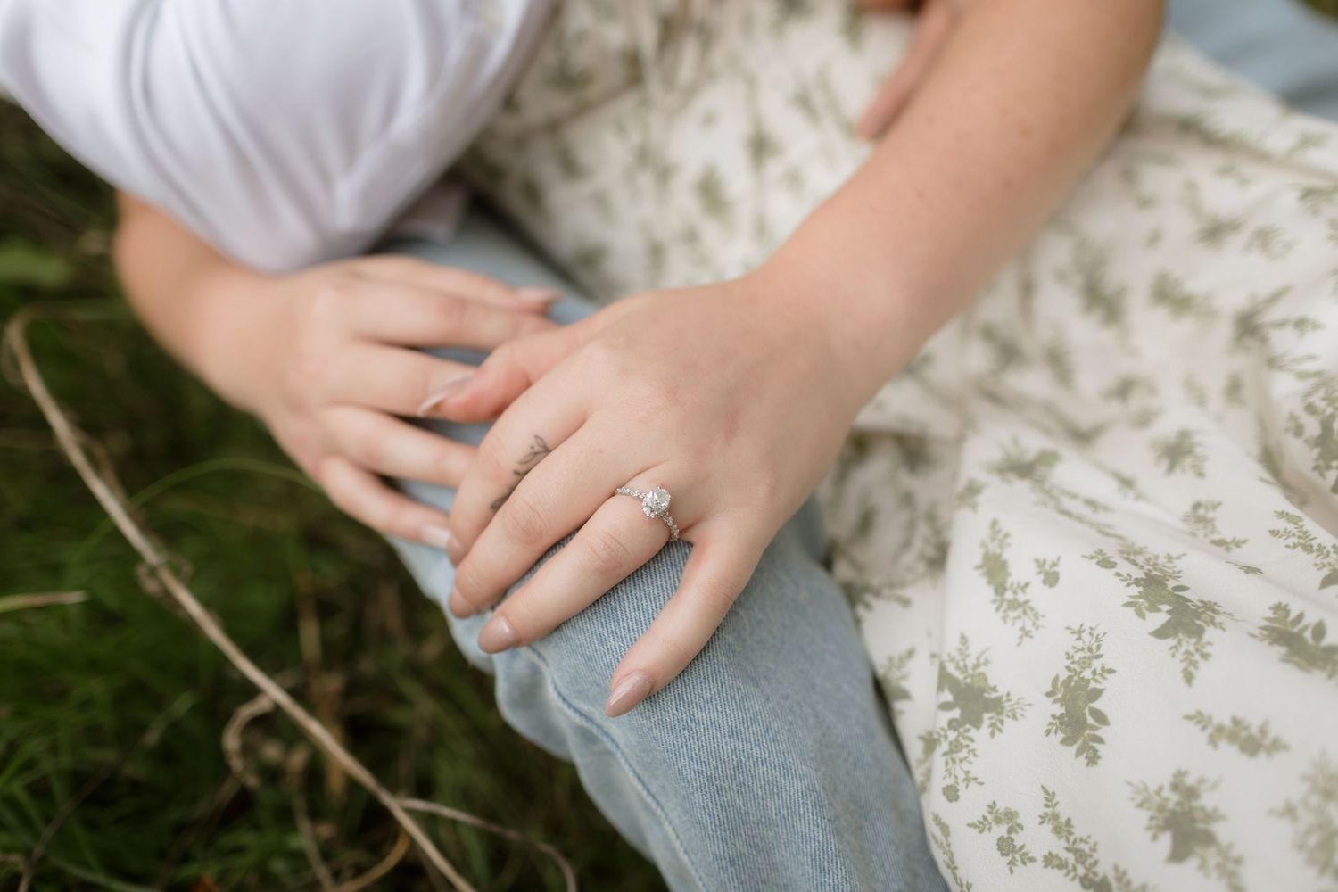 Hand wearing engagement ring rests on knee while sitting on floral patterned fabric outdoors.