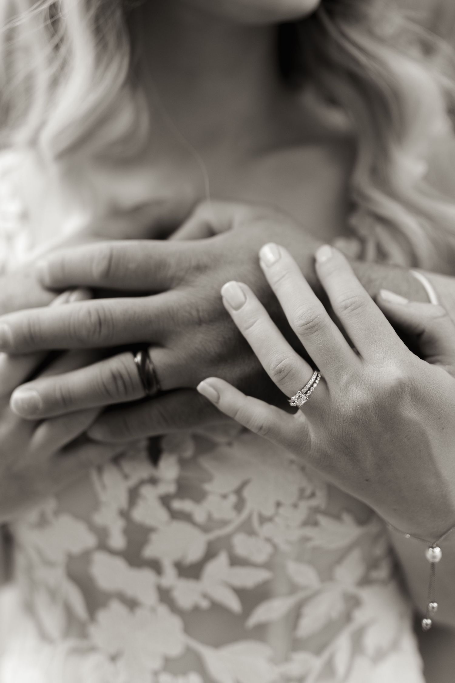 Close-up black and white photo of hands with wedding ring against lace dress fabric.