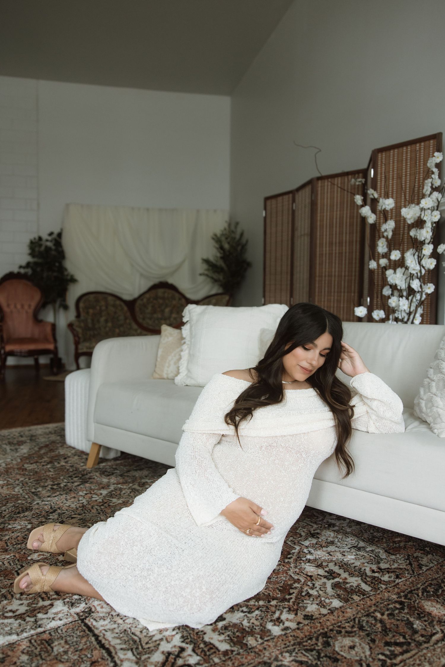 Artistic maternity portrait on patterned rug with white furniture and floral decor.