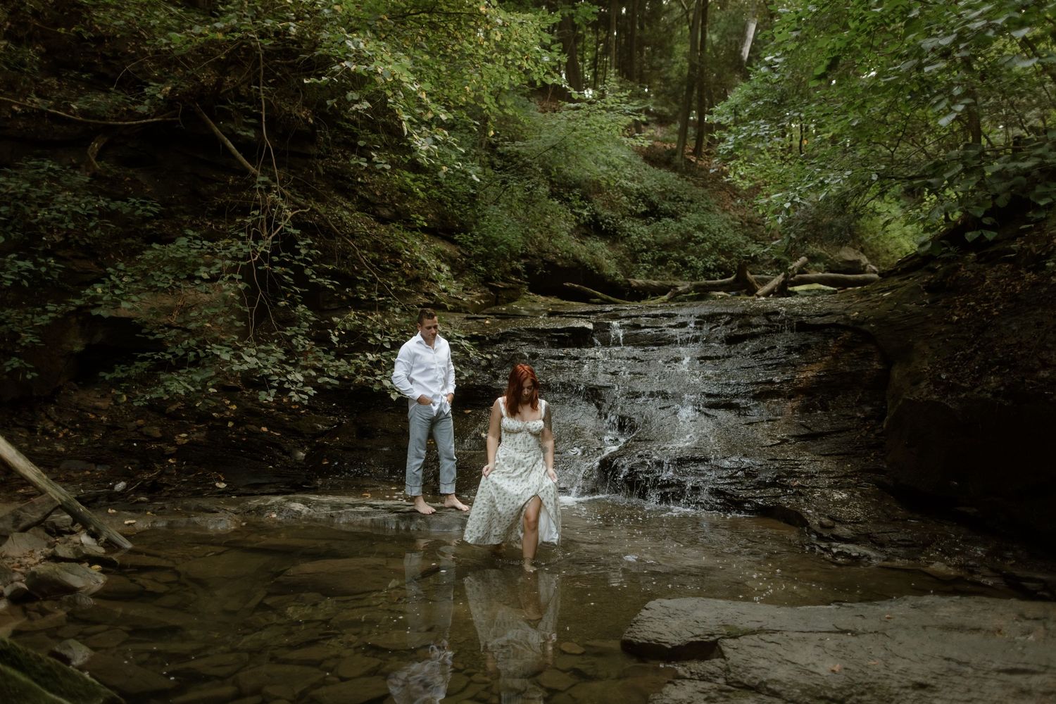 A couple stands in front of a cascading waterfall in a lush forest creek, wearing casual white attire.