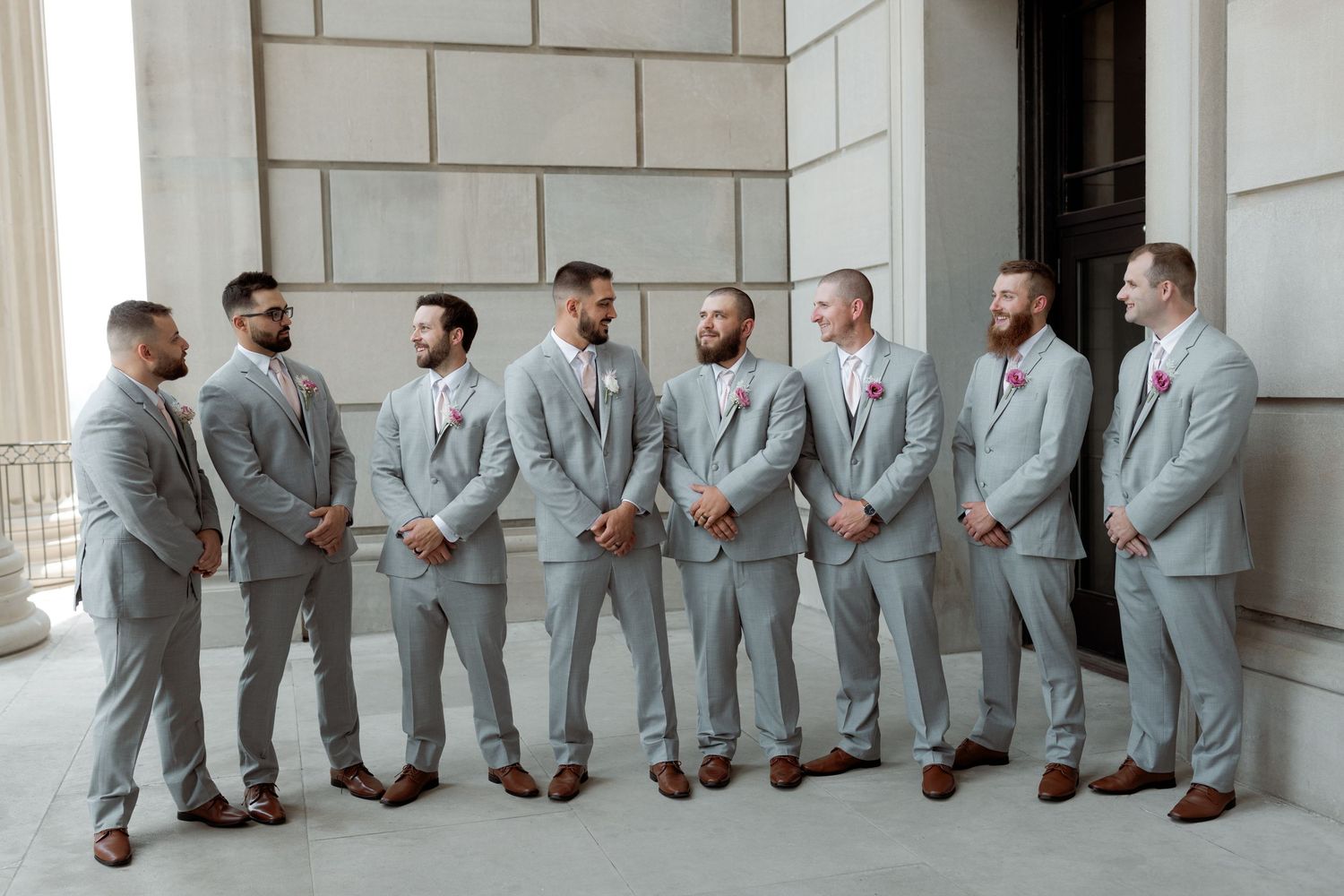 Wedding party of groomsmen in matching light gray suits and brown shoes lined up against stone wall.