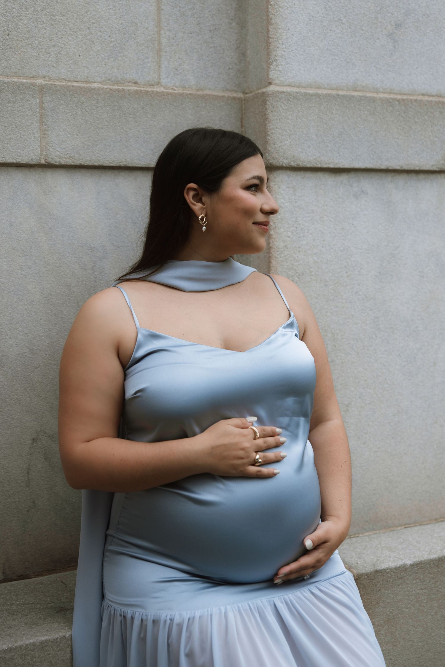 A pregnant woman in a flowing blue dress poses against a stone wall with a gentle smile.