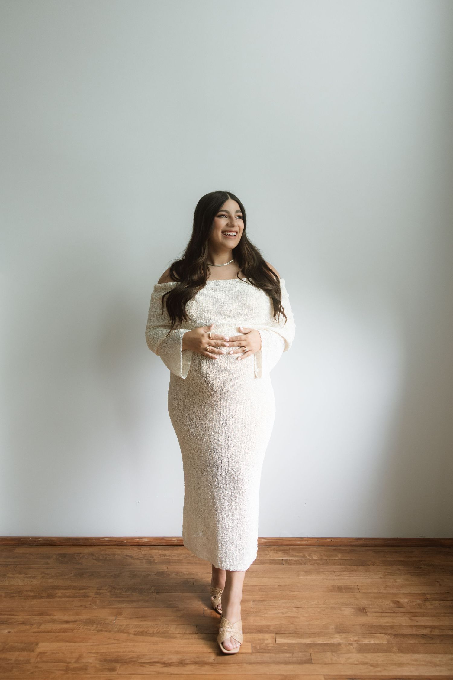 Someone wearing a white lace dress poses playfully in a sequence of minimalist maternity photos against a light wall.
