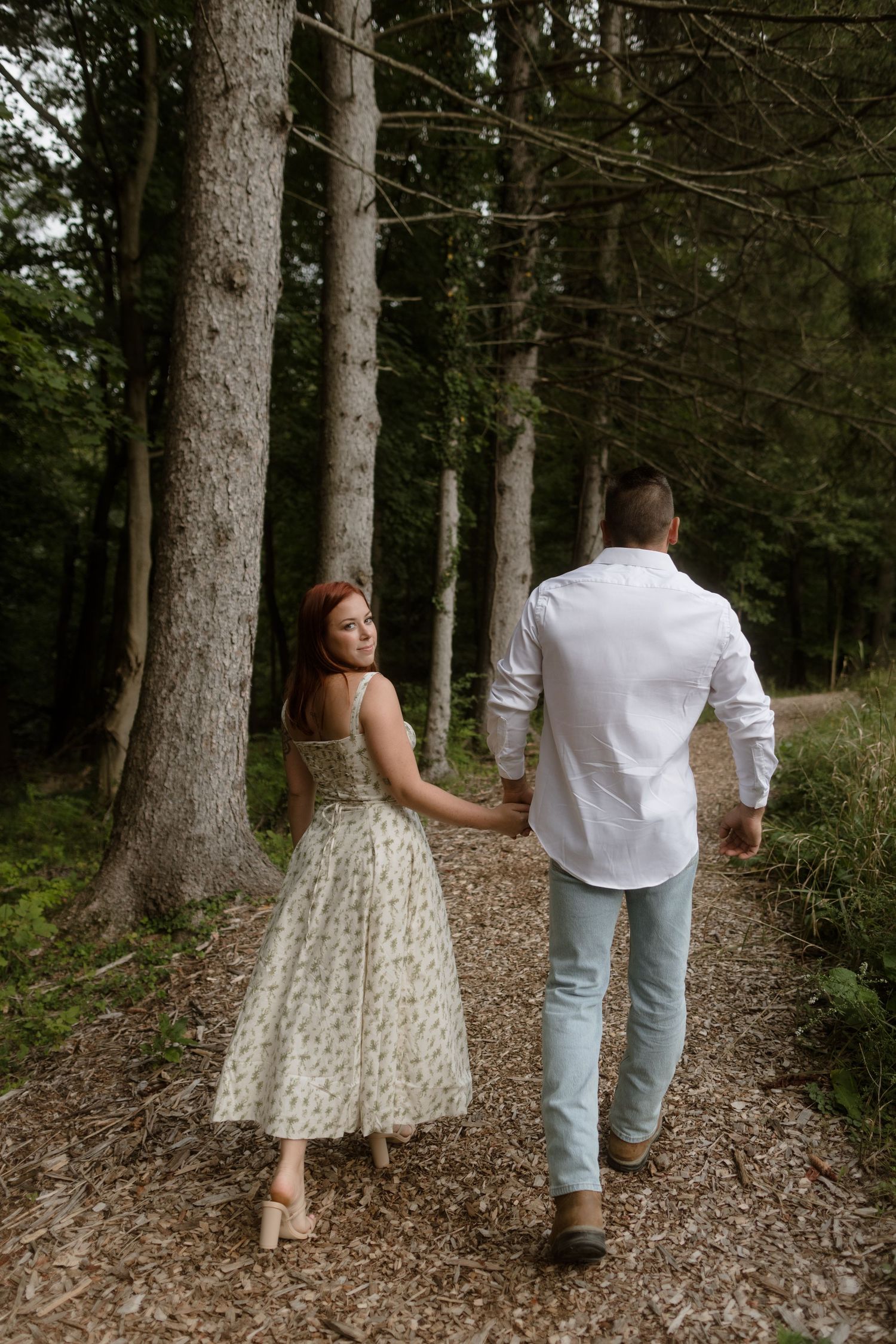 A couple walks hand in hand down a wooded forest path, the woman wearing a polka dot tea-length dress.