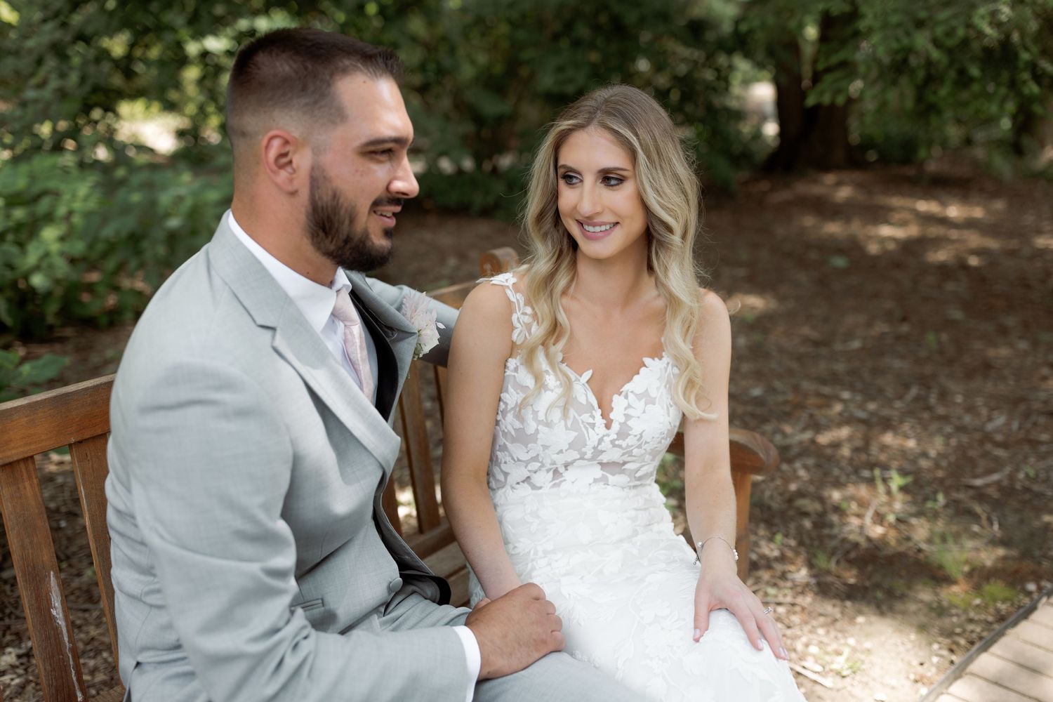 Couple in wedding attire sitting together on wooden bench in outdoor garden setting.