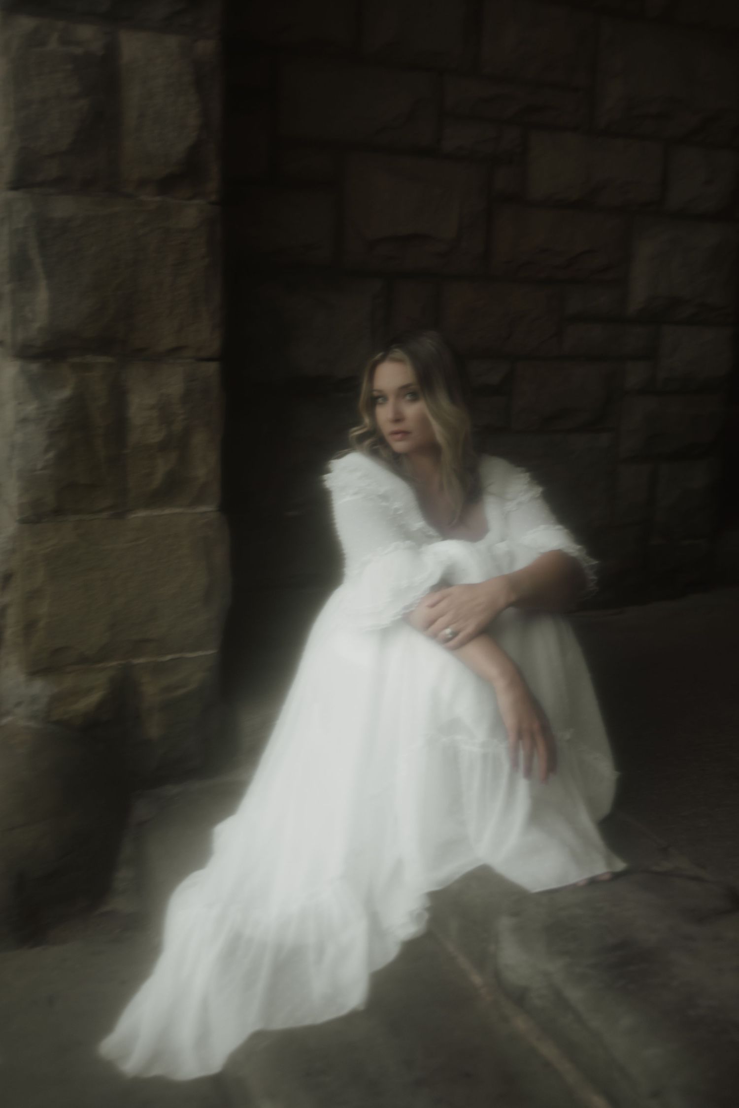 A figure in a flowing white dress sits dreamily against a stone wall in a moody, ethereal portrait.