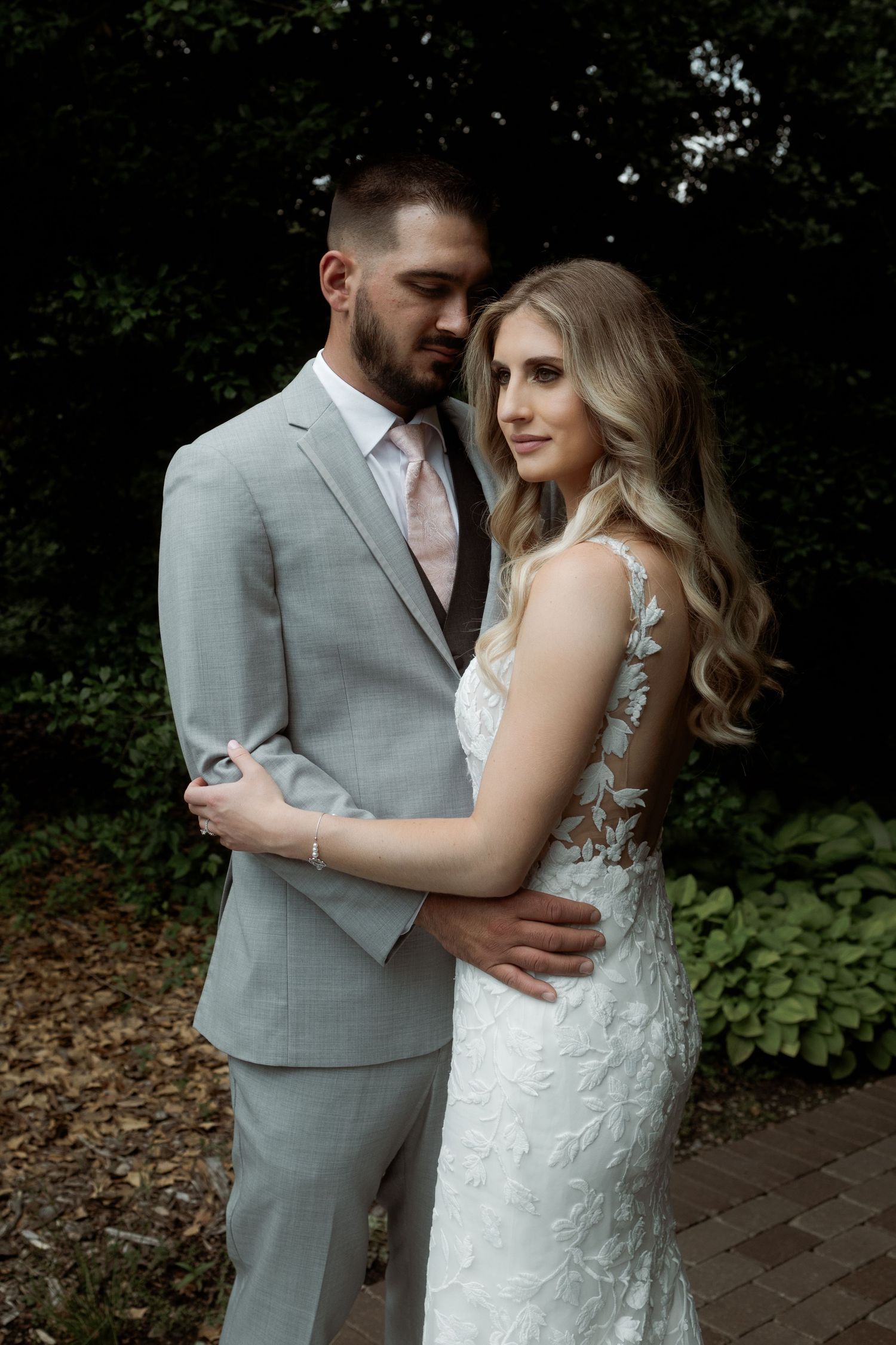 Newlyweds in gray suit and lace wedding dress pose intimately against lush garden backdrop.
