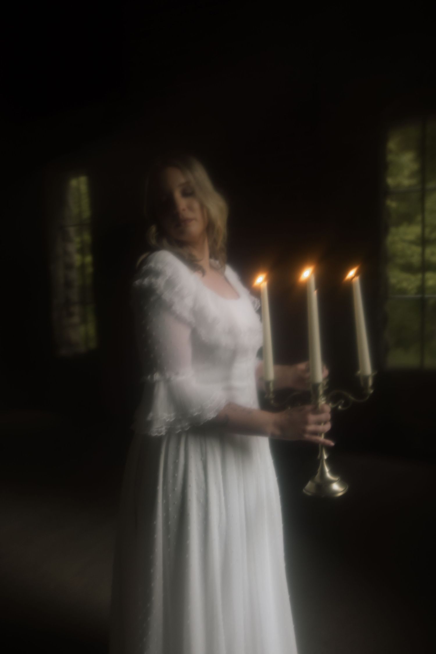 Figure in flowing white dress holds glowing candelabra in atmospheric dark setting with soft focus lighting effects.