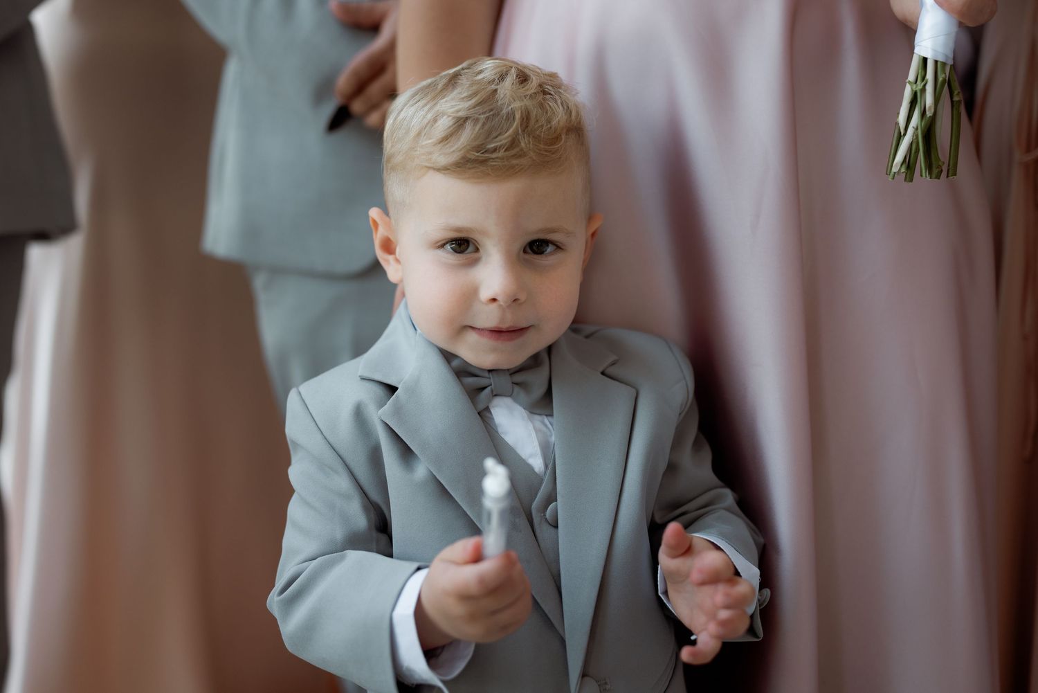 Young ring bearer in light gray suit smiles at camera during wedding ceremony.