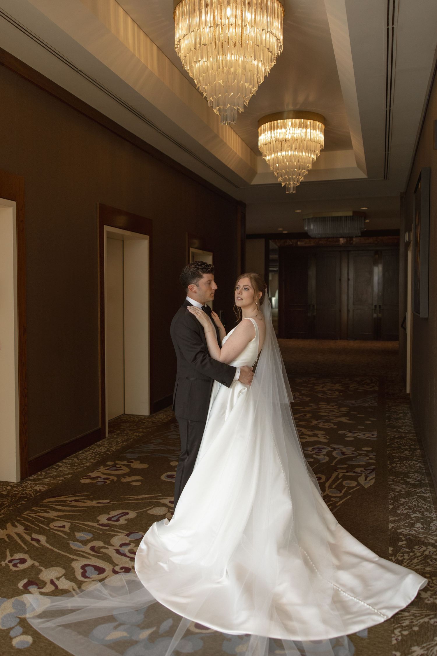 Wedding couple dancing under crystal chandeliers in an elegant hotel hallway.