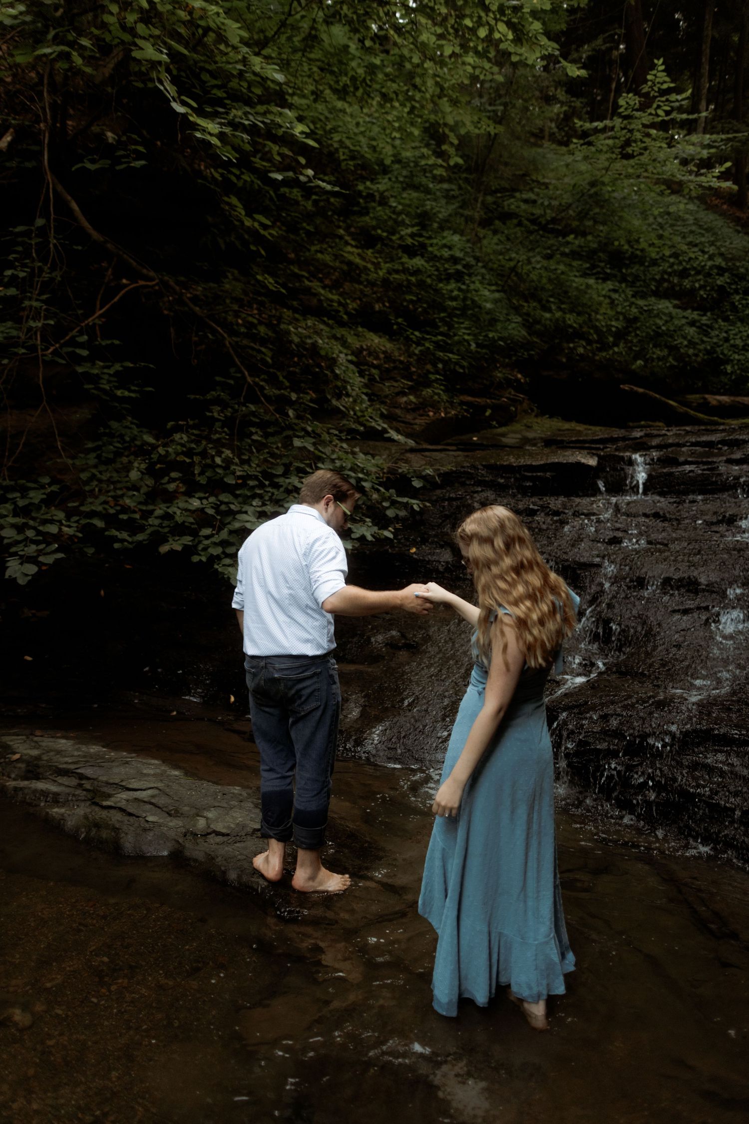 Two people stand together in a shallow creek surrounded by rocky walls and lush forest greenery.