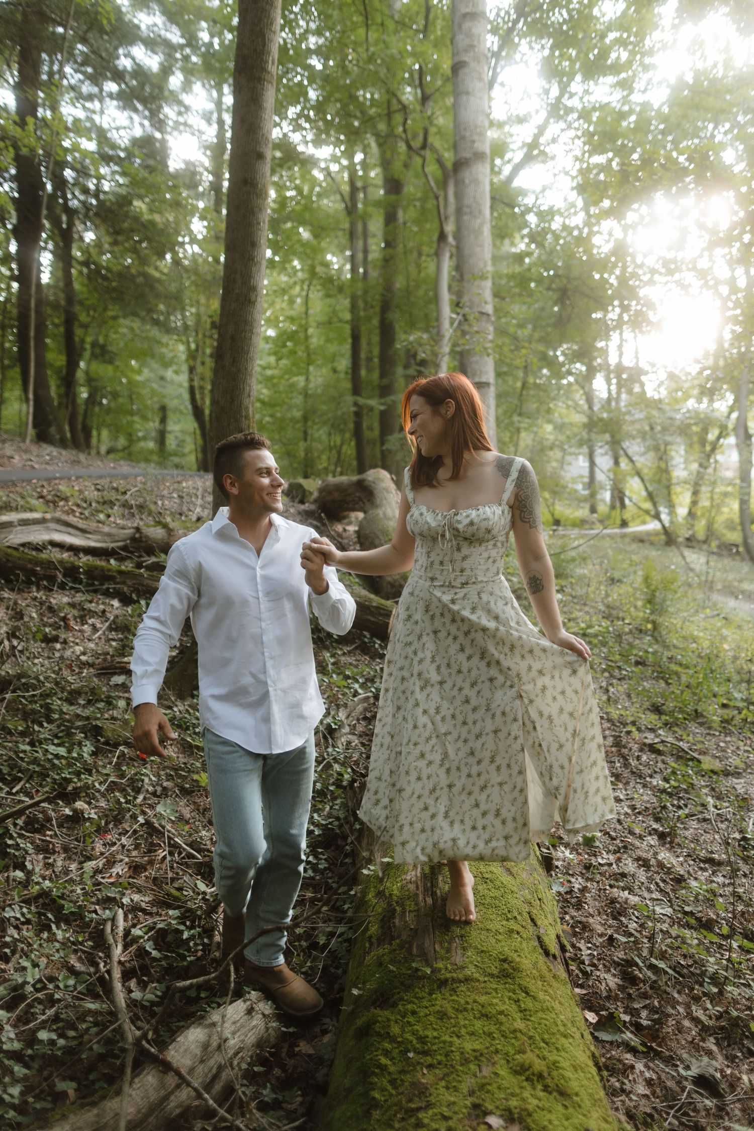 Couple in light summer attire sharing a joyful moment together on a moss-covered log in a sunlit forest.