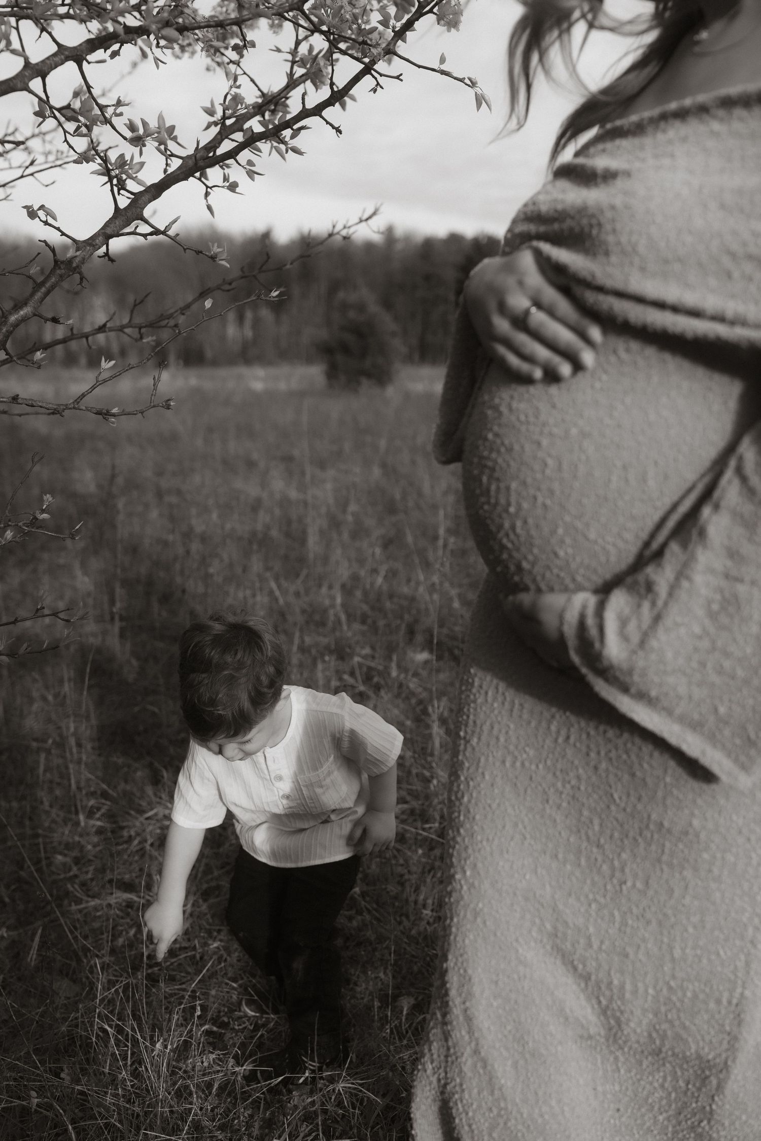 Black and white photo of child playing near flowering trees with person in background.