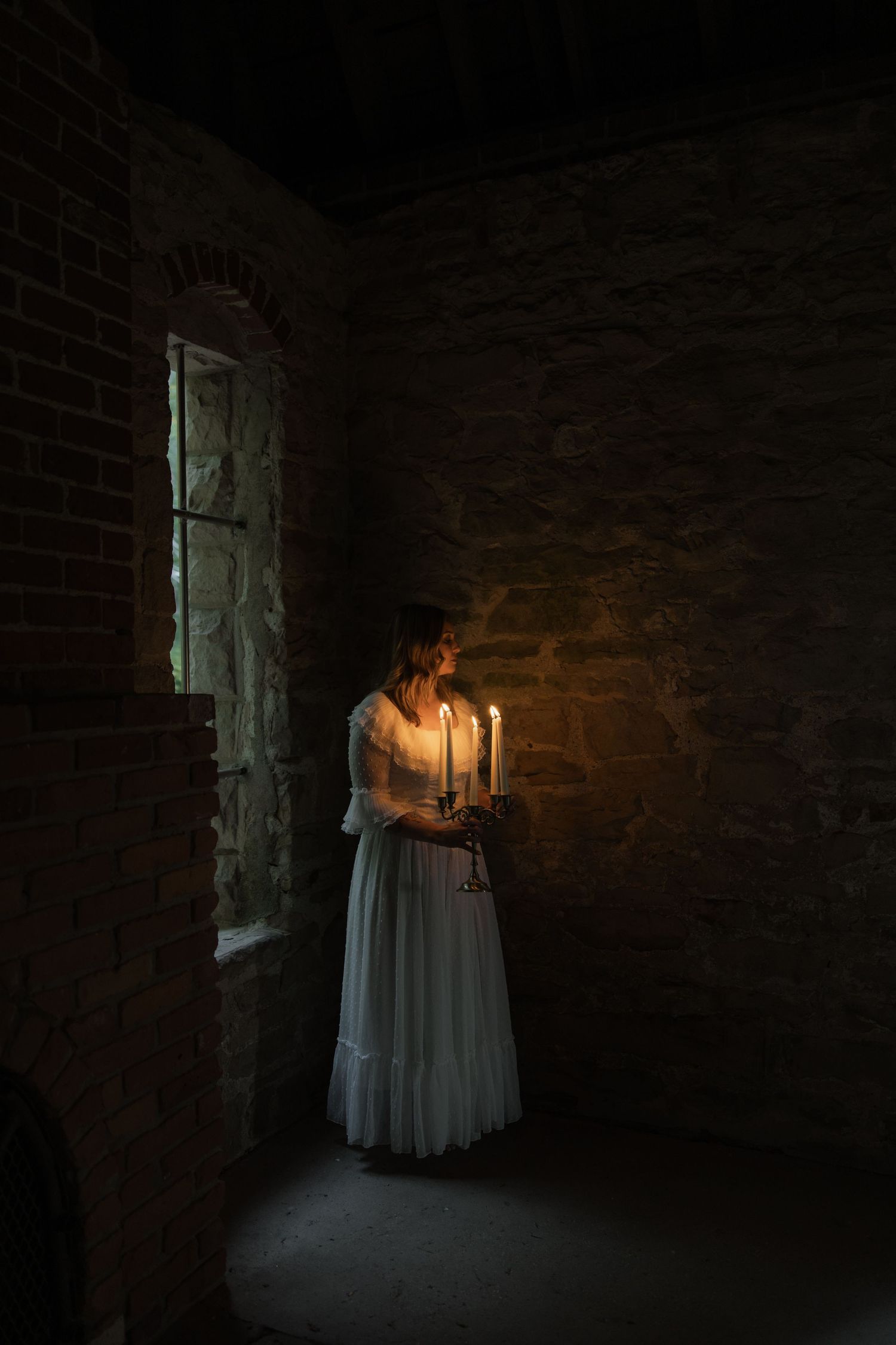 A figure in a white dress holds a candlestick while standing in a dimly lit brick room near a window.