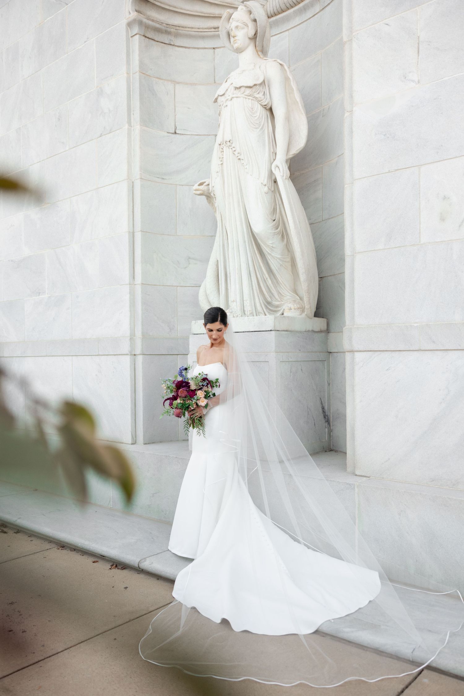 A bride in a classic white wedding gown with a flowing train stands elegantly against a marble wall with classical statues.