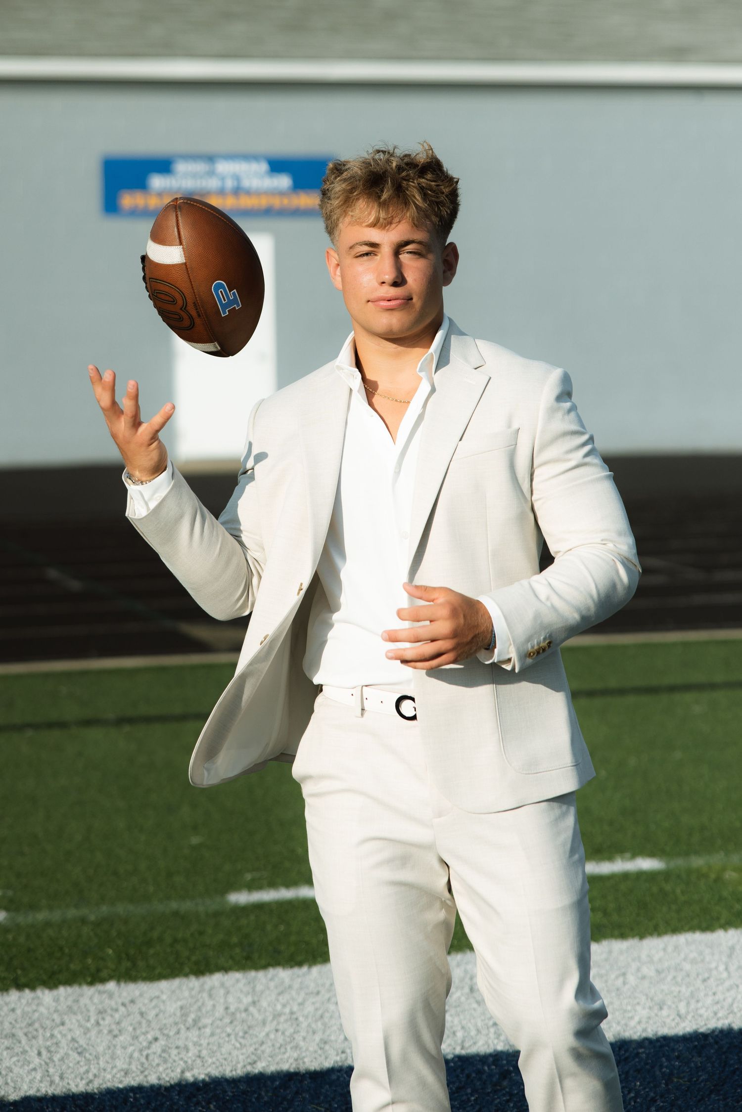 A person in a white suit holding and posing with a football on an athletic field.