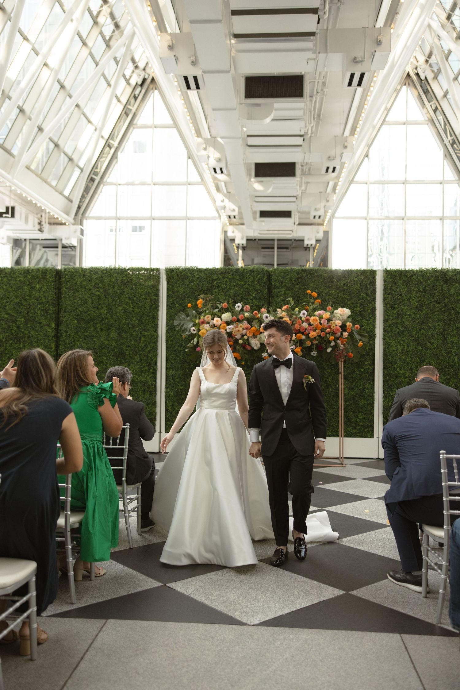 Wedding ceremony processional in a modern greenhouse with dramatic vaulted glass ceiling.