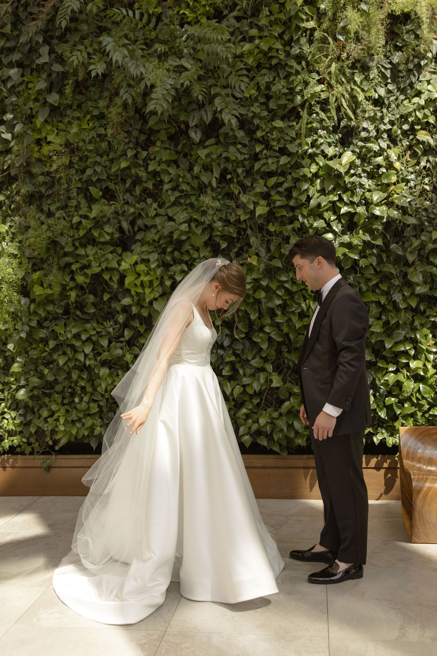 Wedding couple poses against a lush green ivy wall backdrop, bride in elegant white gown with long train.