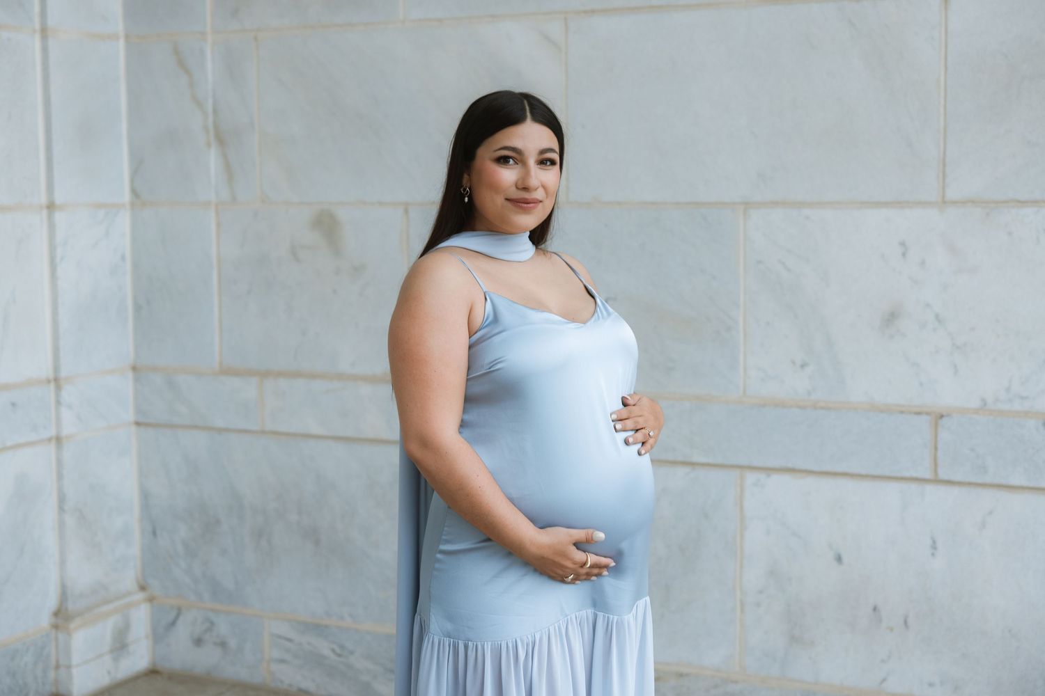 Expectant woman in a light blue maxi dress poses against a marble wall outdoors.
