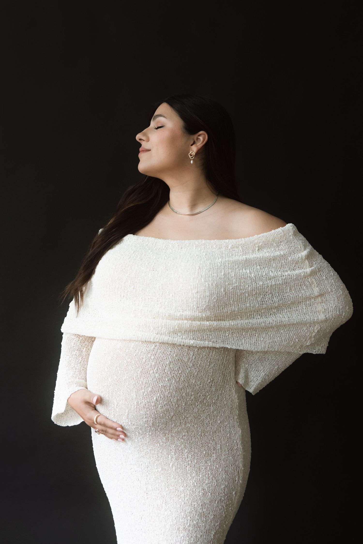 A graceful maternity photo features a woman in an elegant off-shoulder white sequined gown against a dark background.