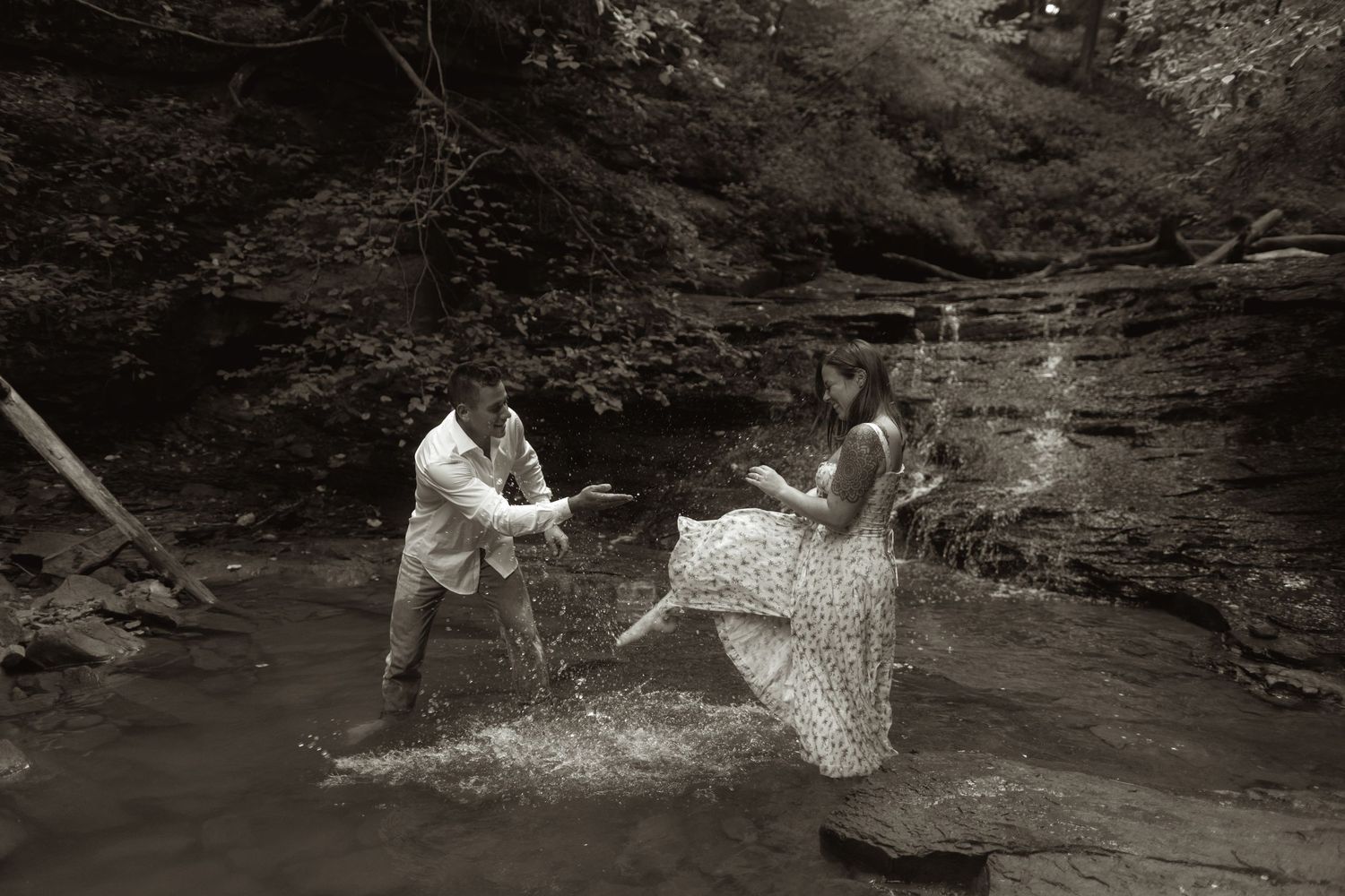 Two people creating a splashing moment in front of a waterfall in a serene forest setting, captured in black and white.