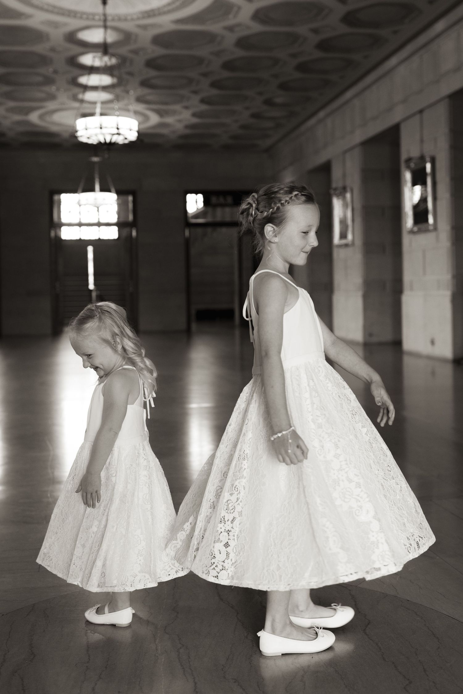 Two young flower girls in white lace dresses stand side by side in an elegant hallway with ornate ceiling lights.