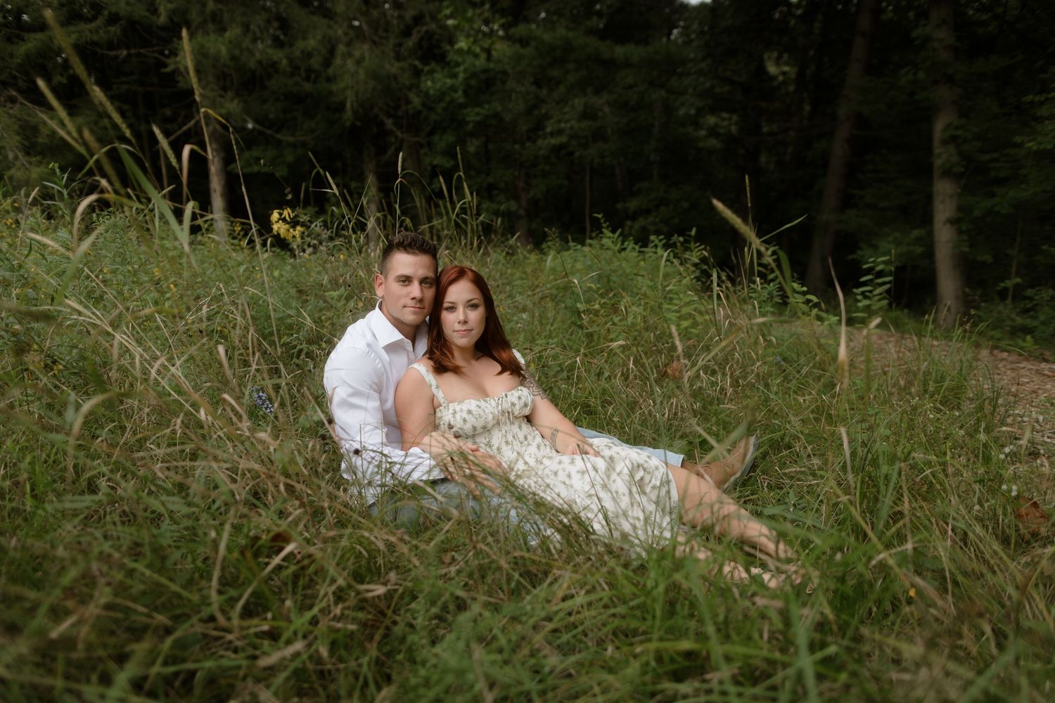 Two people resting together in a romantic pose among tall wild grasses at the edge of a wooded area.