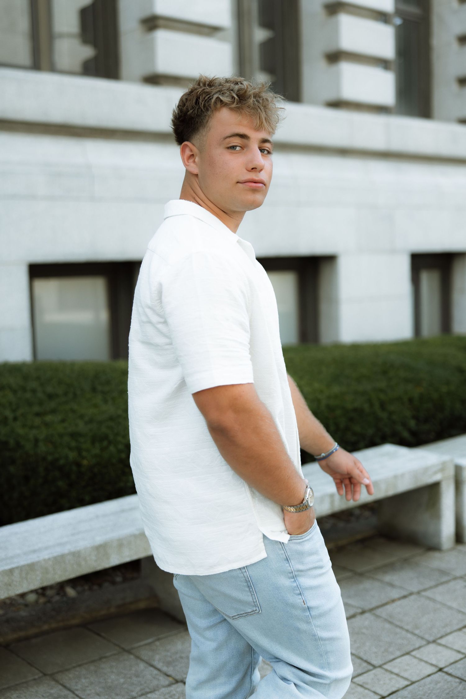 Person wearing white shirt and light blue pants poses casually against outdoor wall and benches.