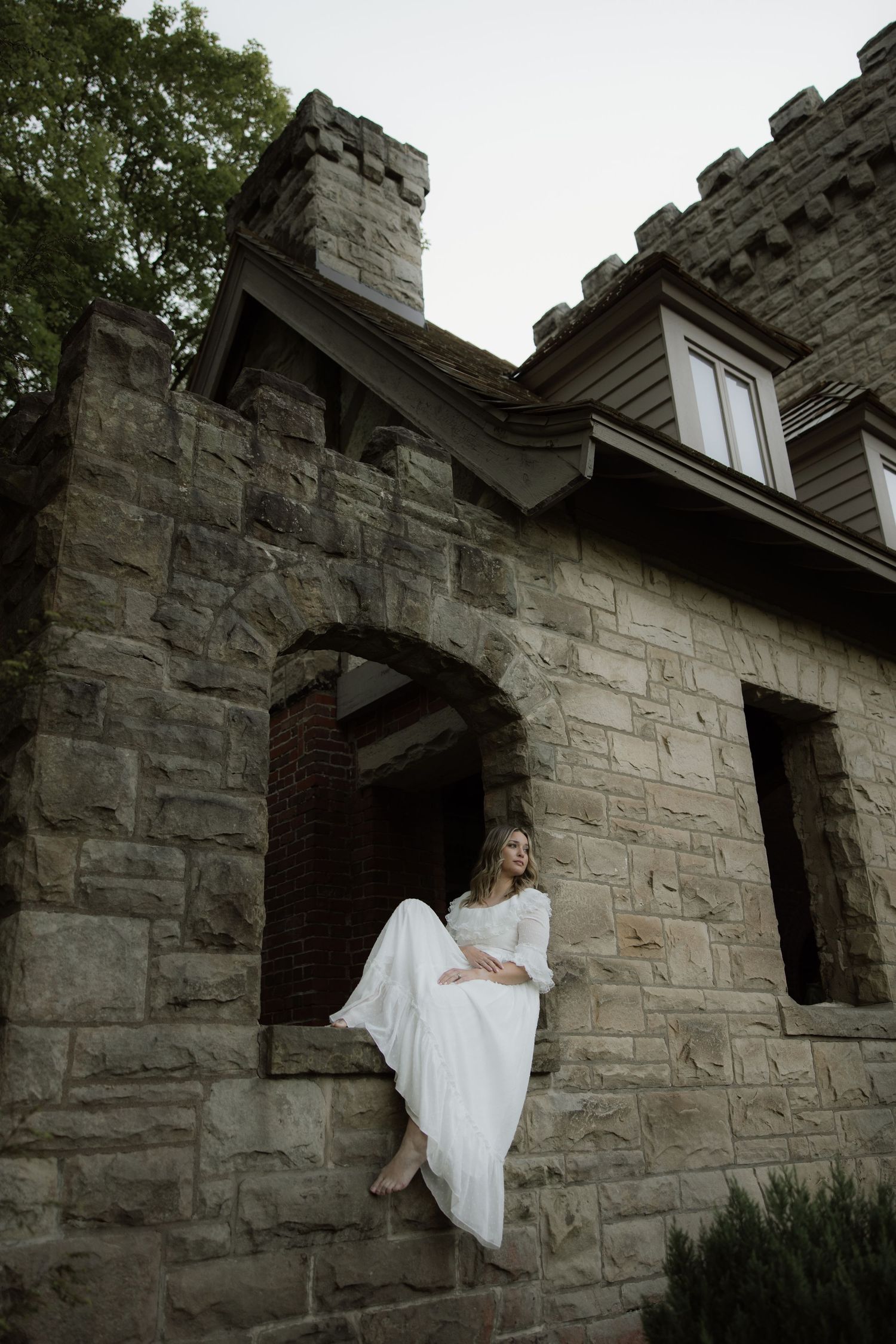 Figure in flowing white dress sits perched on stone archway of historic castle-like building at dusk.