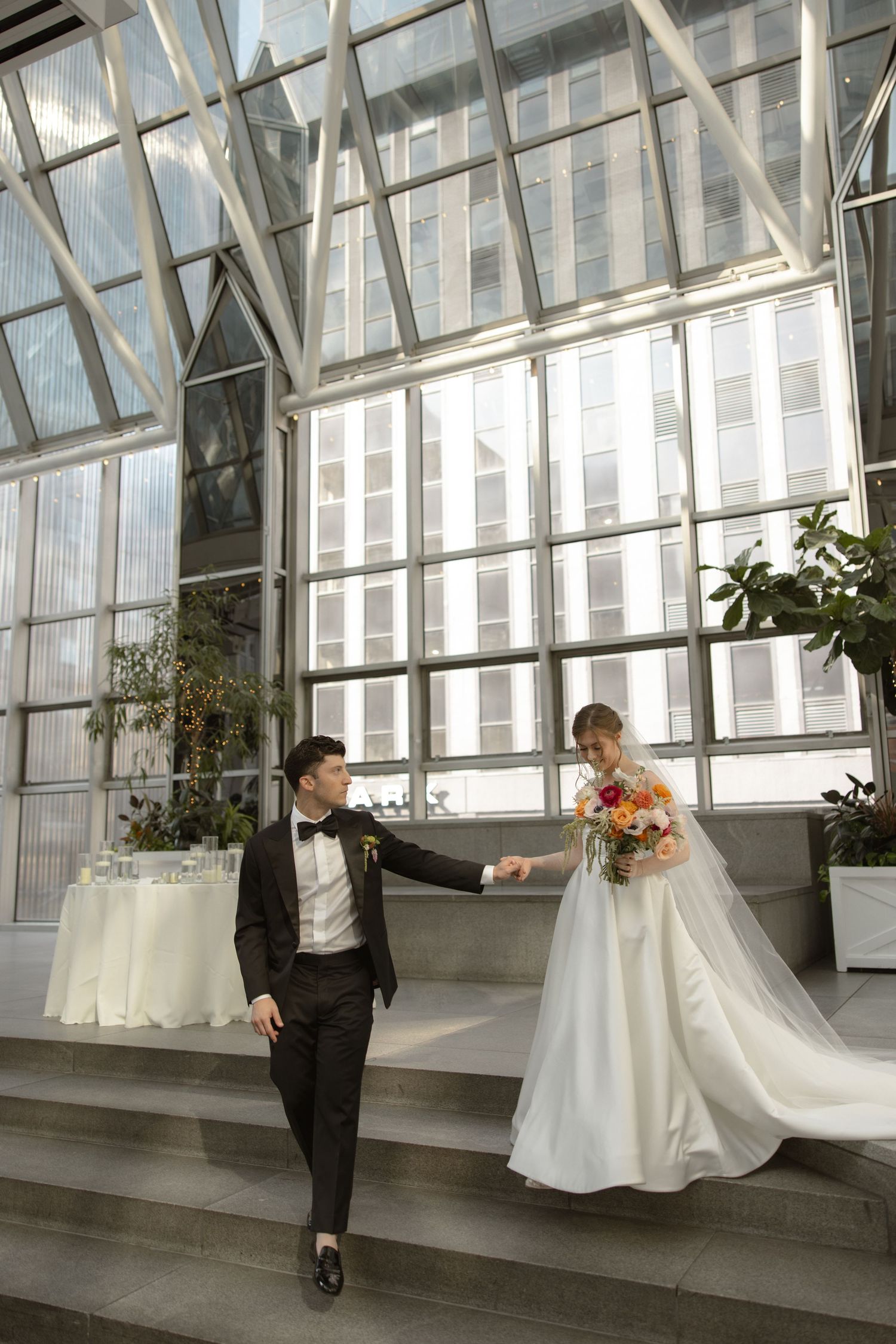Wedding couple descending glass-enclosed stairs, bride holding colorful bouquet.