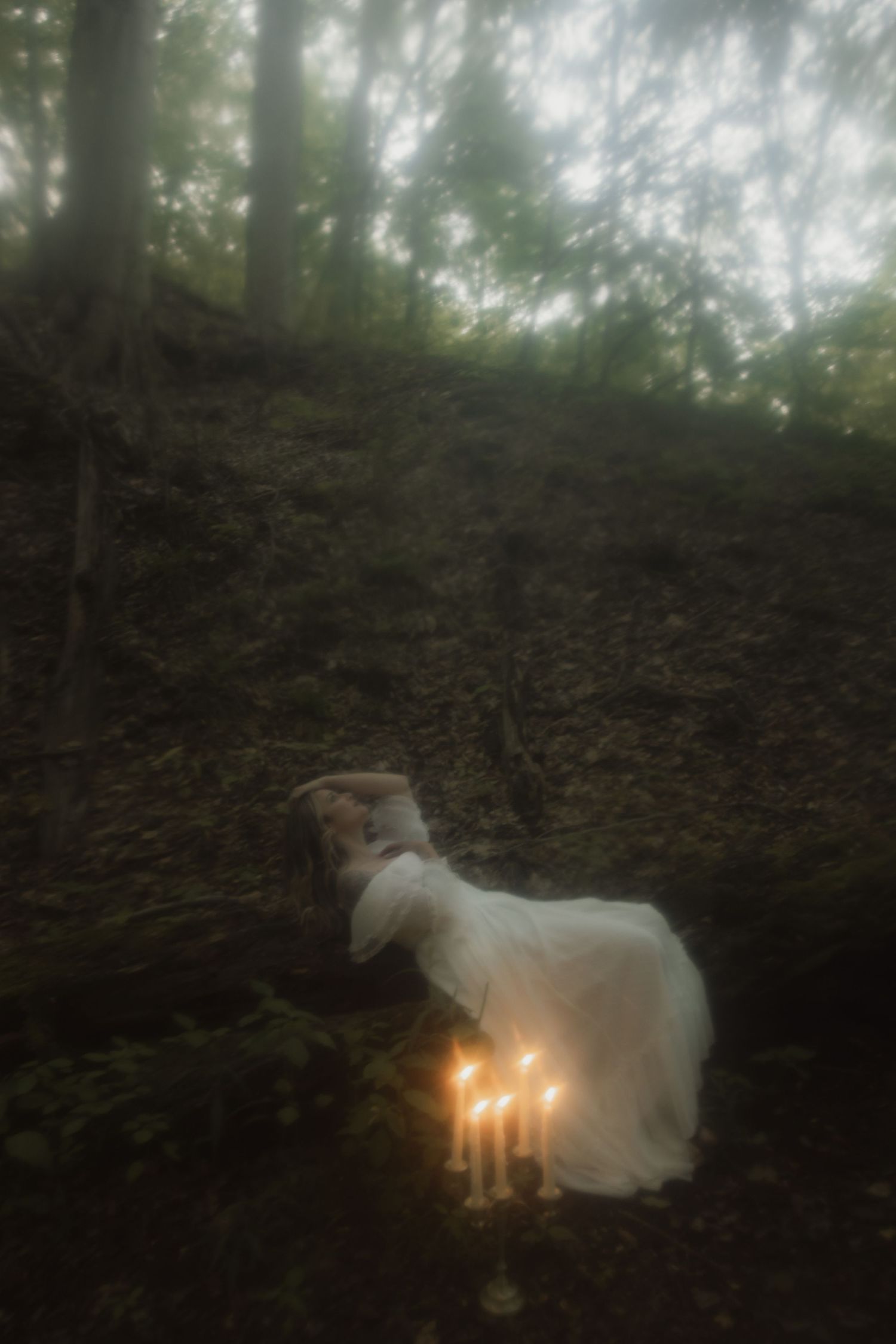 Ethereal figure in white dress rests on forest floor beside glowing candlelight creating mystical woodland atmosphere.