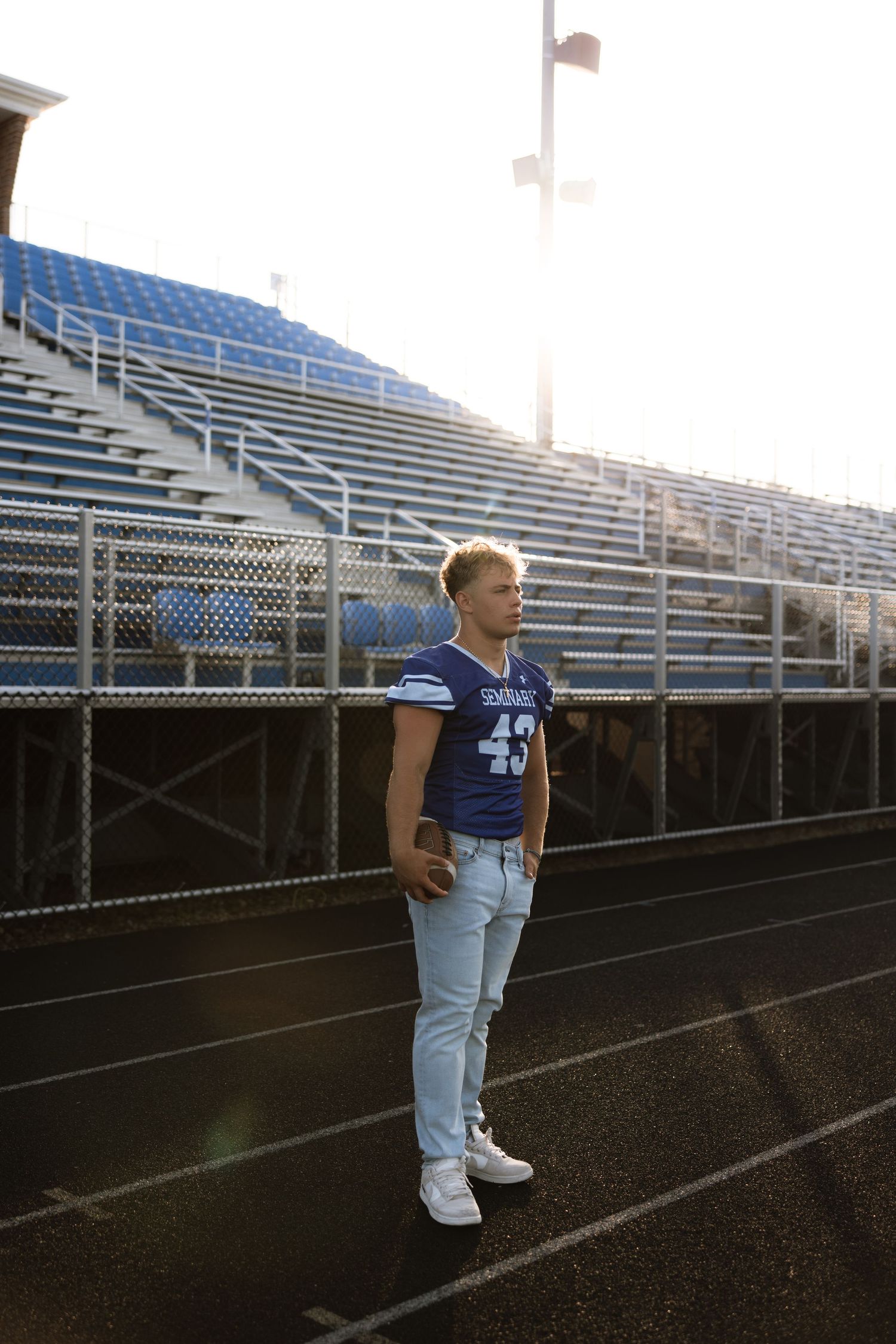 Football player in a varsity jersey stands alone on a track field with empty bleachers in the background at sunset.
