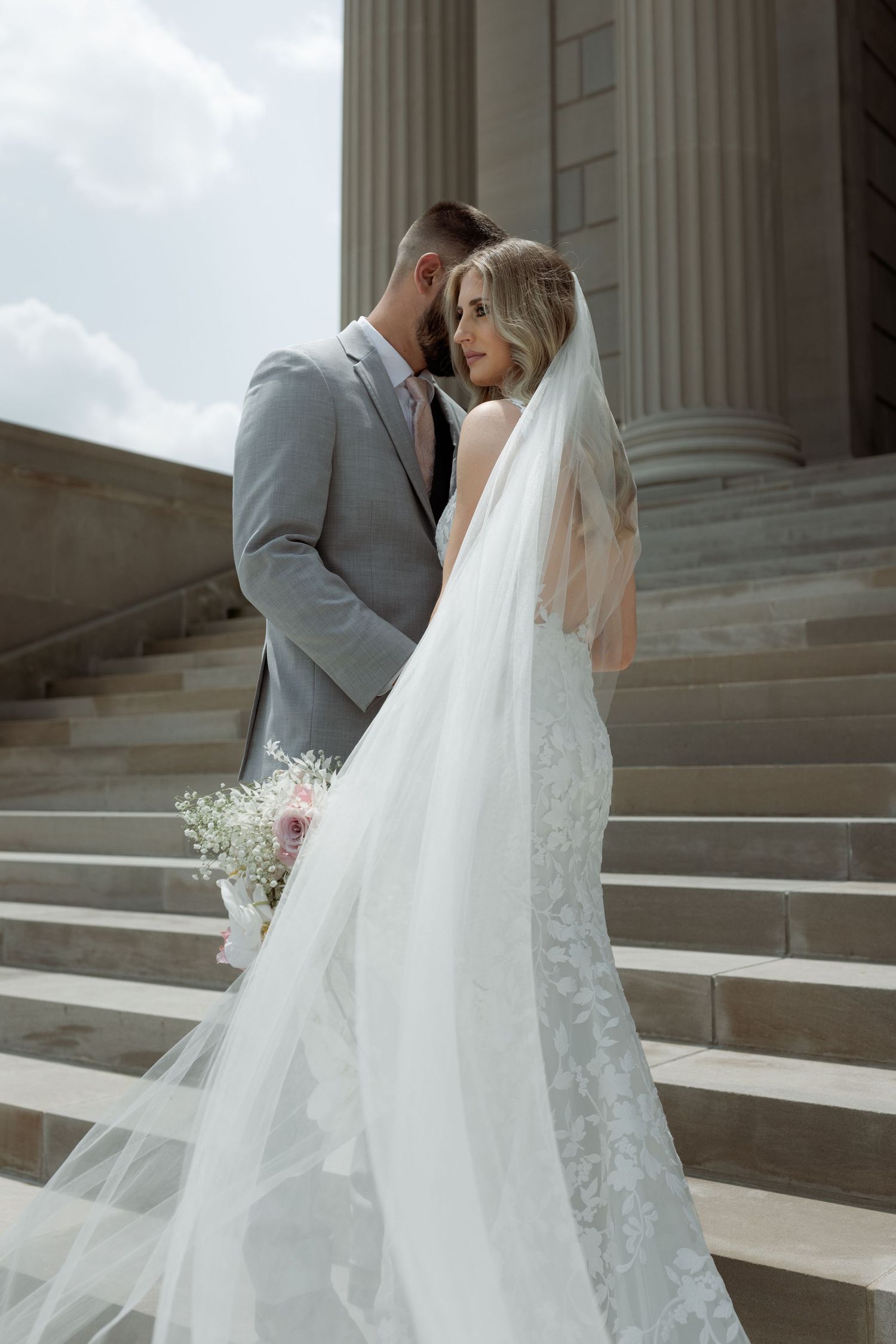 Romantic bride and groom embrace on stone steps, with long flowing veil and gray suit against classical columns.