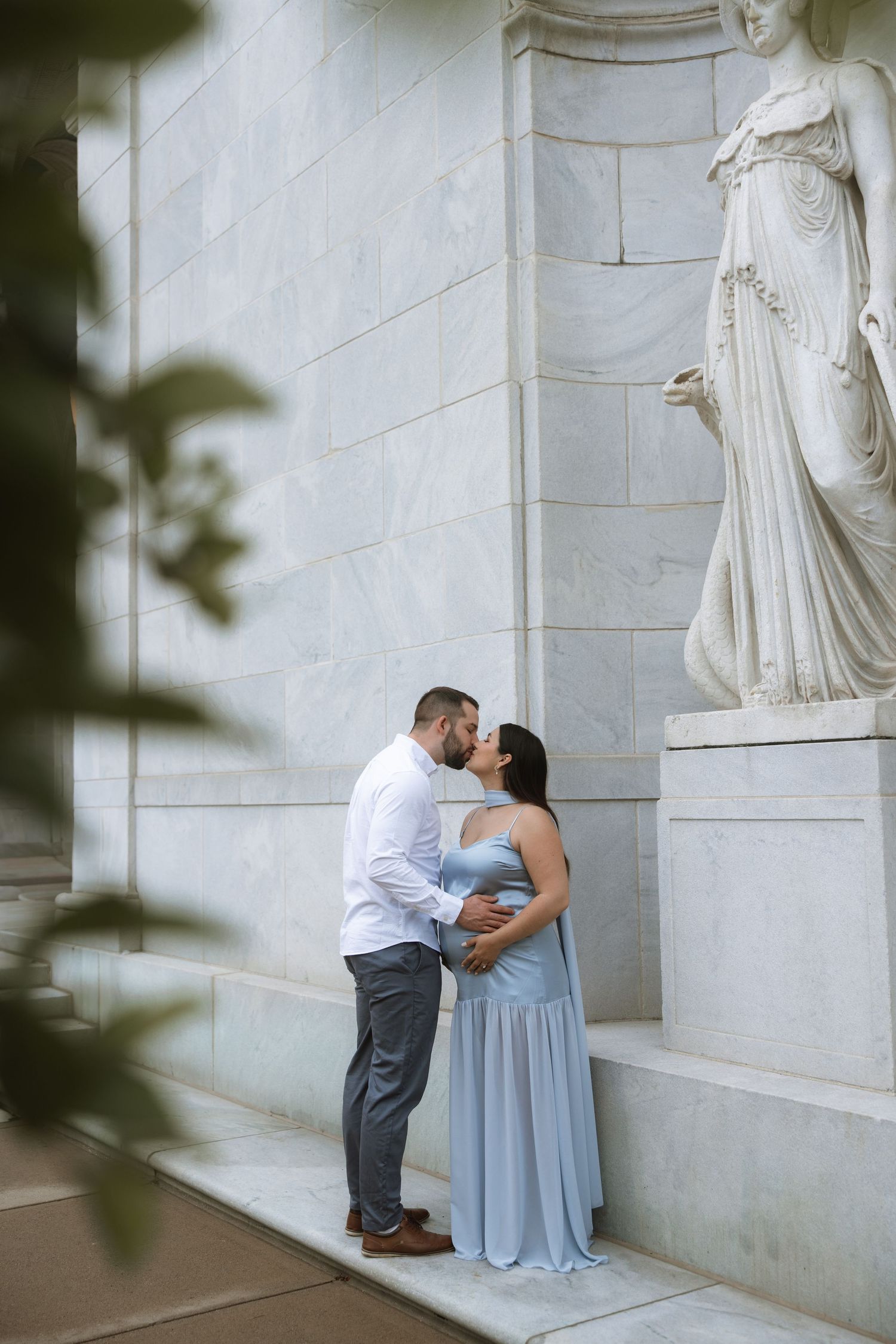 A romantic maternity photo of a couple embracing by a classical stone statue in soft lighting.