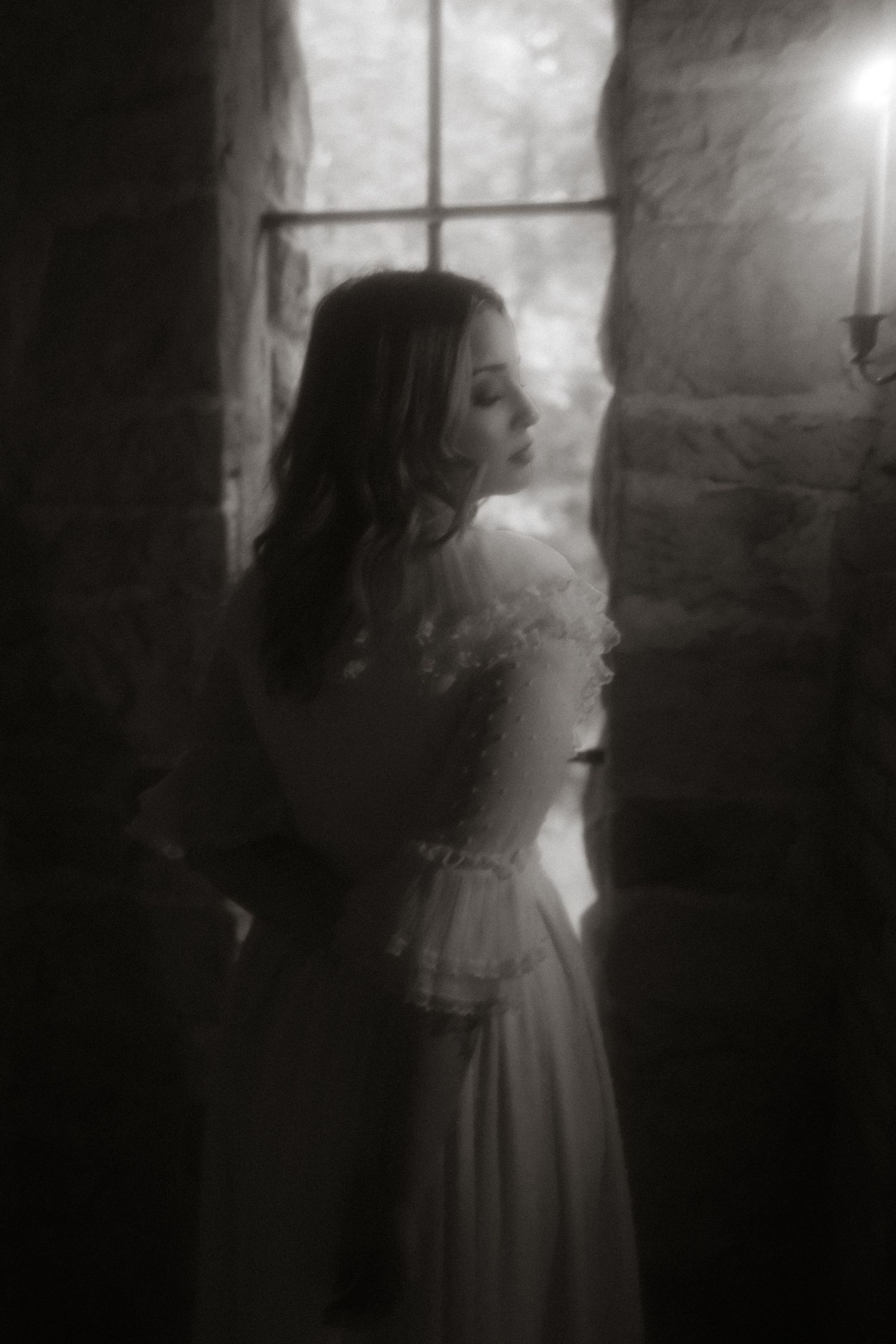 A dreamy black and white portrait of a person in a vintage dress standing by a window in moody atmospheric lighting.