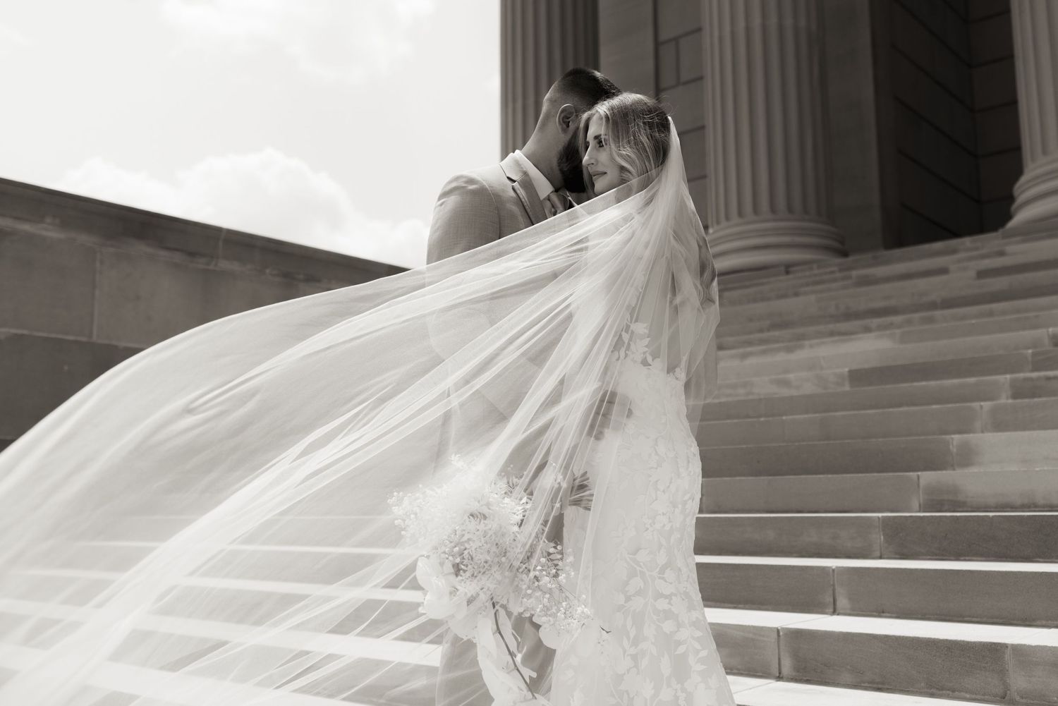 Flowing wedding veil catches the breeze on stone steps of a classical building.