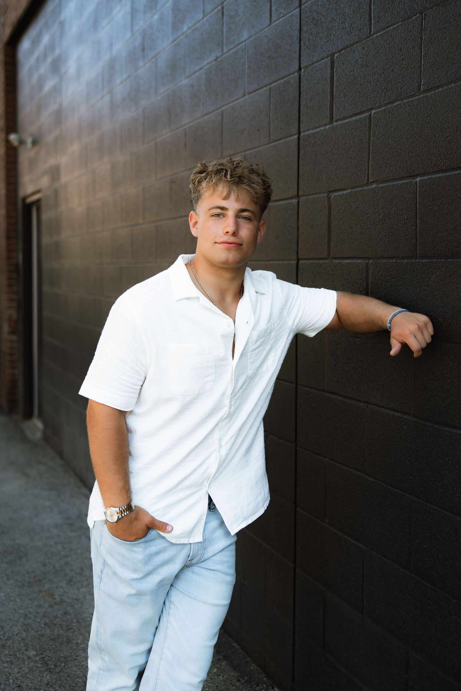 A person in a white shirt and light-colored pants poses casually against a dark wall in an outdoor setting.