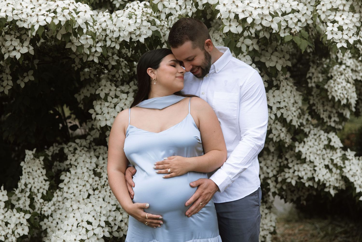 A couple embraces in front of a blooming white floral backdrop during a maternity photoshoot.