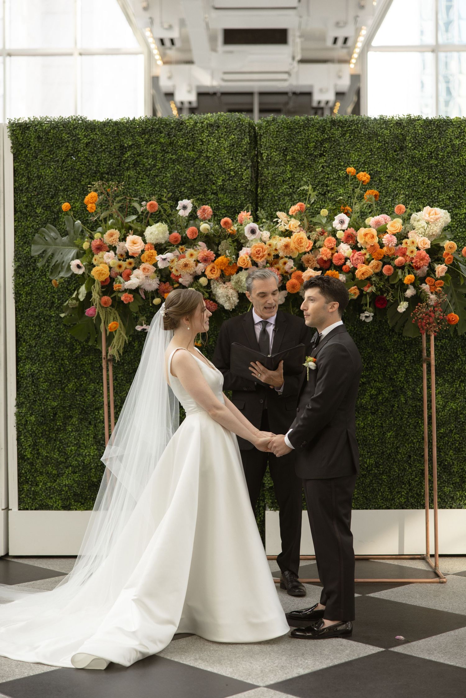 Elegant wedding ceremony setup with a floral arch featuring orange and white blooms against a greenery wall.