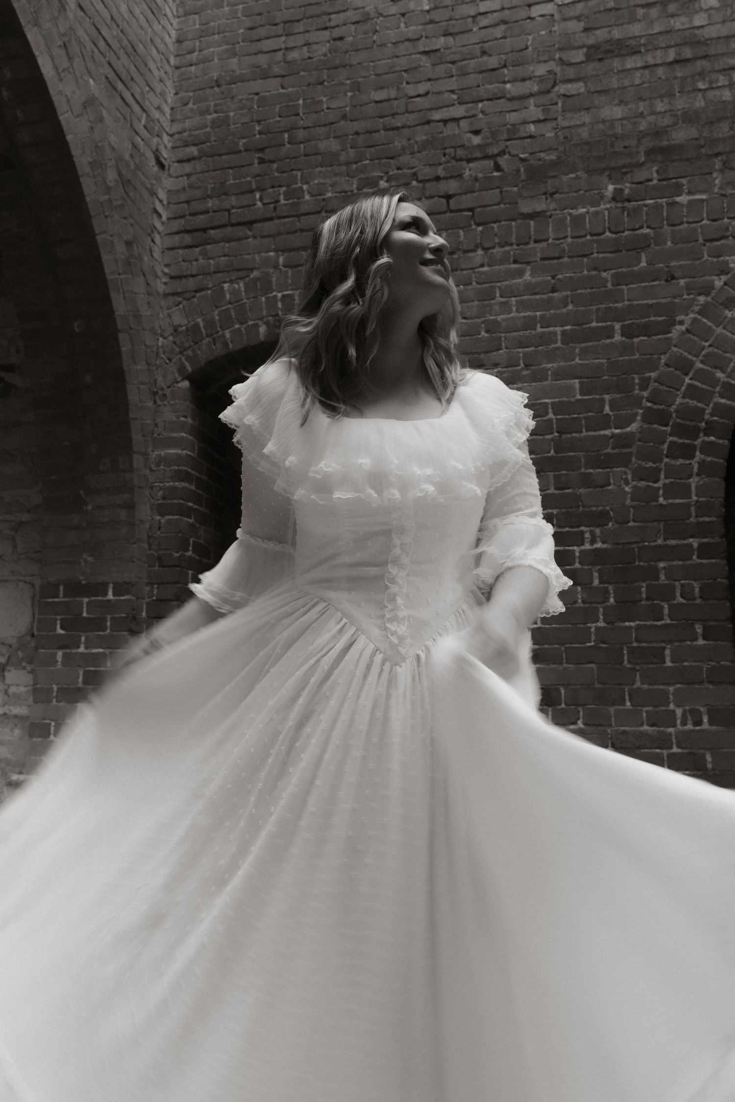 A vintage-style black and white photo showing a twirling wedding dress with ruffled details against a brick wall background.