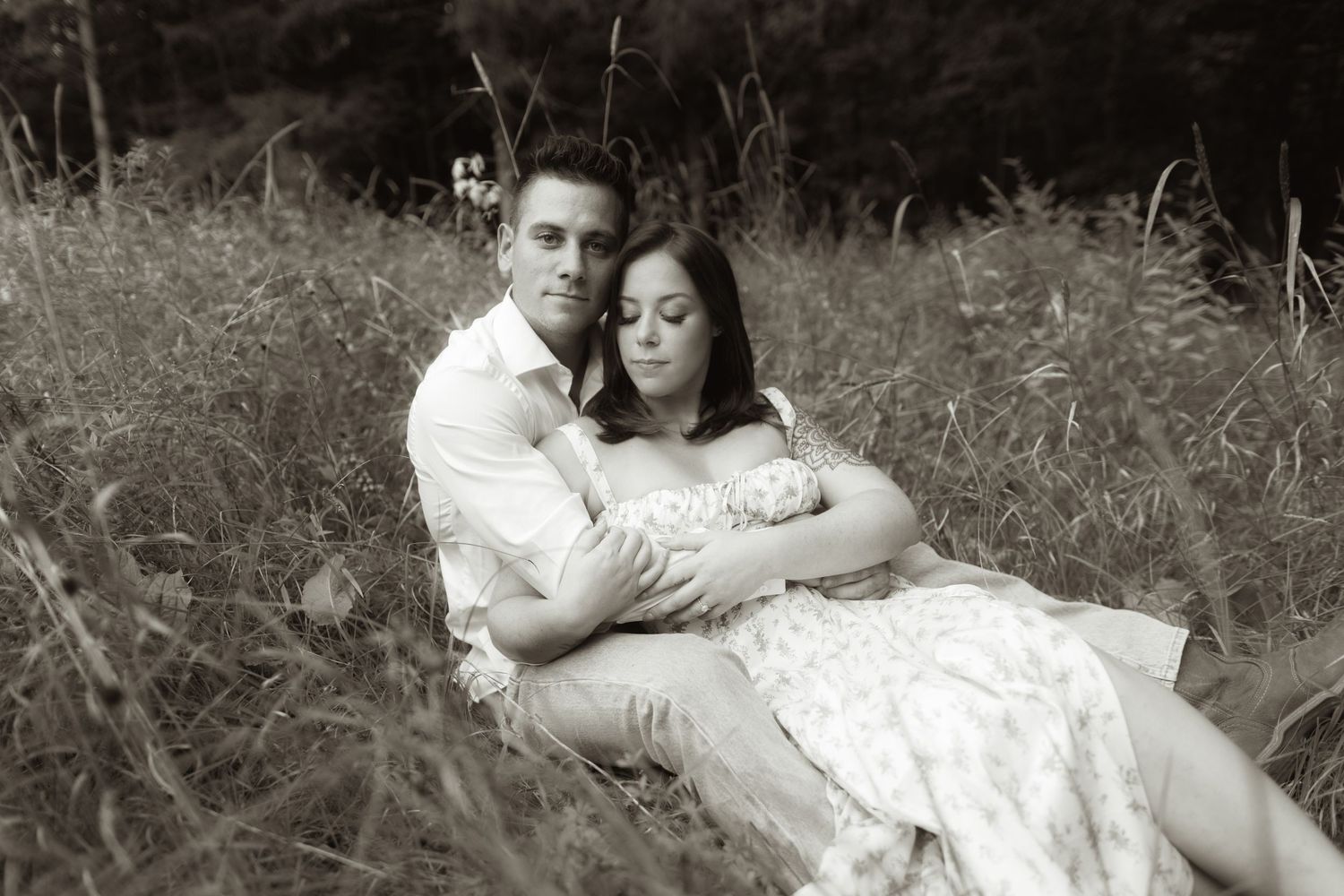 A romantic black and white portrait of a couple embracing while sitting in a rustic field of tall grass.