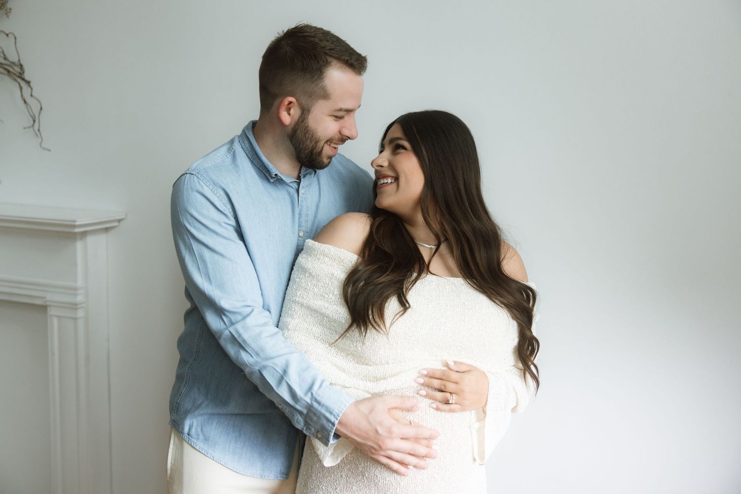 Loving couple embraces intimately while standing against white wall during indoor maternity photo session.