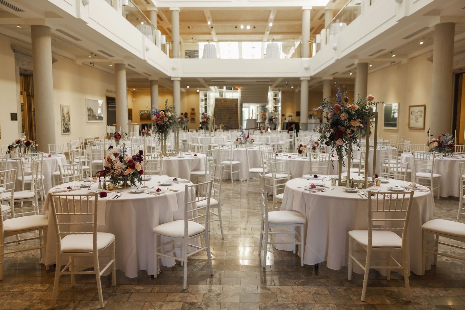Elegant white banquet hall with round tables, chiavari chairs and high ceilings showcasing modern interior design.